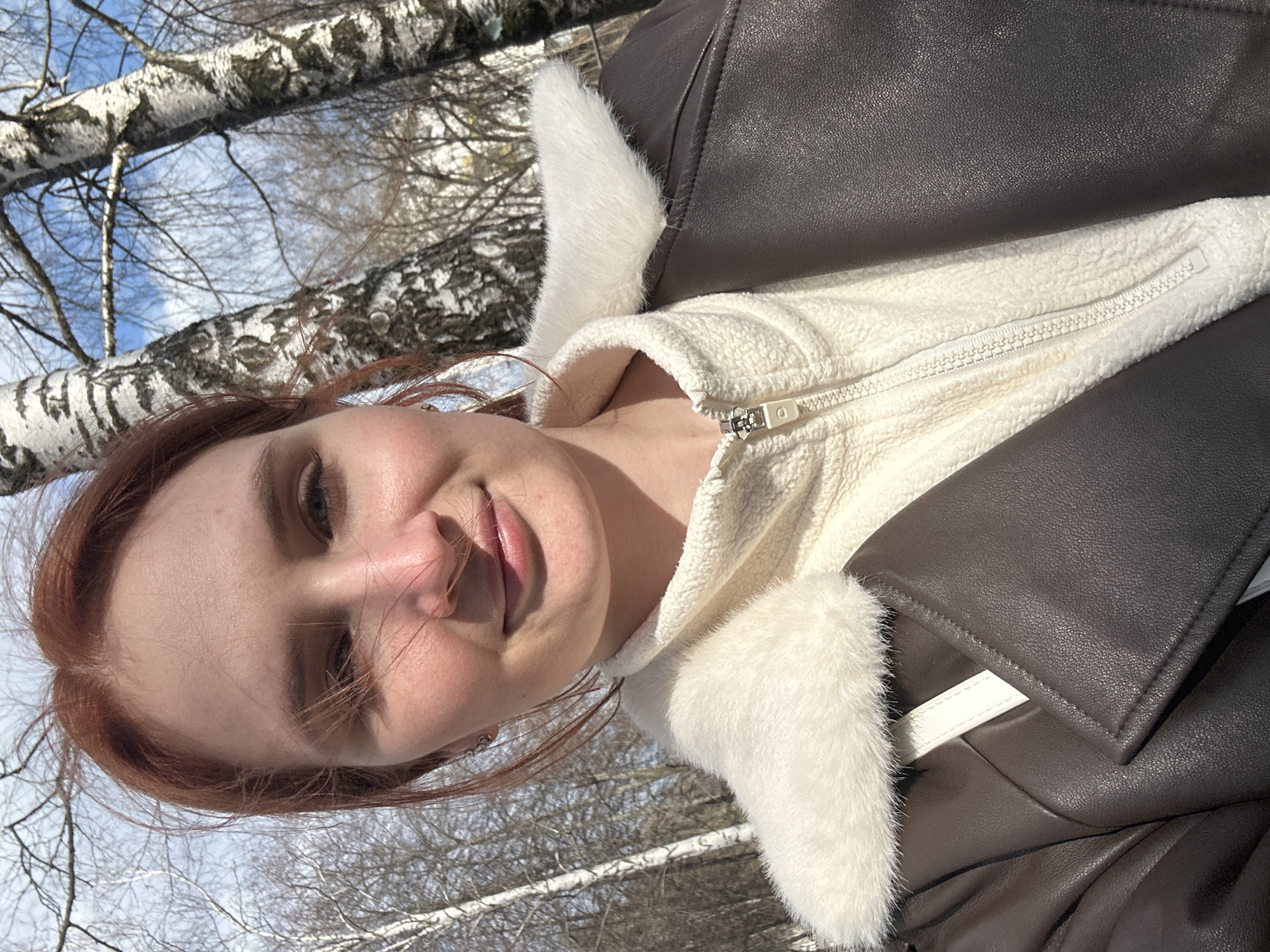 A rotated, close-up selfie captures a young woman outdoors in a birch forest, likely in Chaikovsky, Russia. She looks directly at the camera with a soft, gentle smile, her reddish-brown hair subtly framing her face. Her fair skin is illuminated by bright sunlight.
She is dressed in warm winter or early spring attire, featuring a dark brown faux leather jacket with a plush white faux fur collar and lining. Beneath the jacket, a textured cream or off-white fleece top is visible, fastened with a white zipper that has a matching white pull.
The background consists of a clear blue sky with sparse white clouds, seen through the bare, distinctive white and black barked branches of birch trees. The absence of leaves on the trees indicates either winter or early spring. The bright sunlight suggests a clear, crisp day. The overall scene conveys a serene and peaceful outdoor moment. No visible text is present in the image.