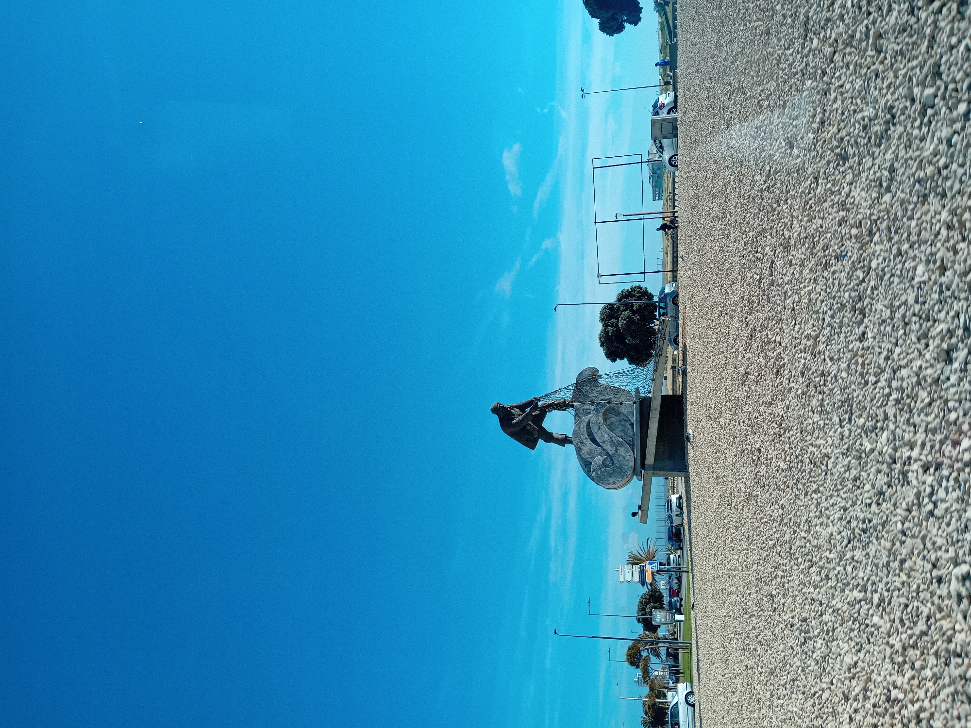 The image captures a side view of the "Monumento ao Homem do Mar" (Monument to the Man of the Sea), a sculpture depicting a fisherman pulling a net. The monument is located in Figueira da Foz, Portugal. It stands on a concrete base along a street with parked cars, trees, and lampposts. A vast expanse of blue sky fills the background, with some light clouds. The ground near the monument is covered with small white stones or gravel.