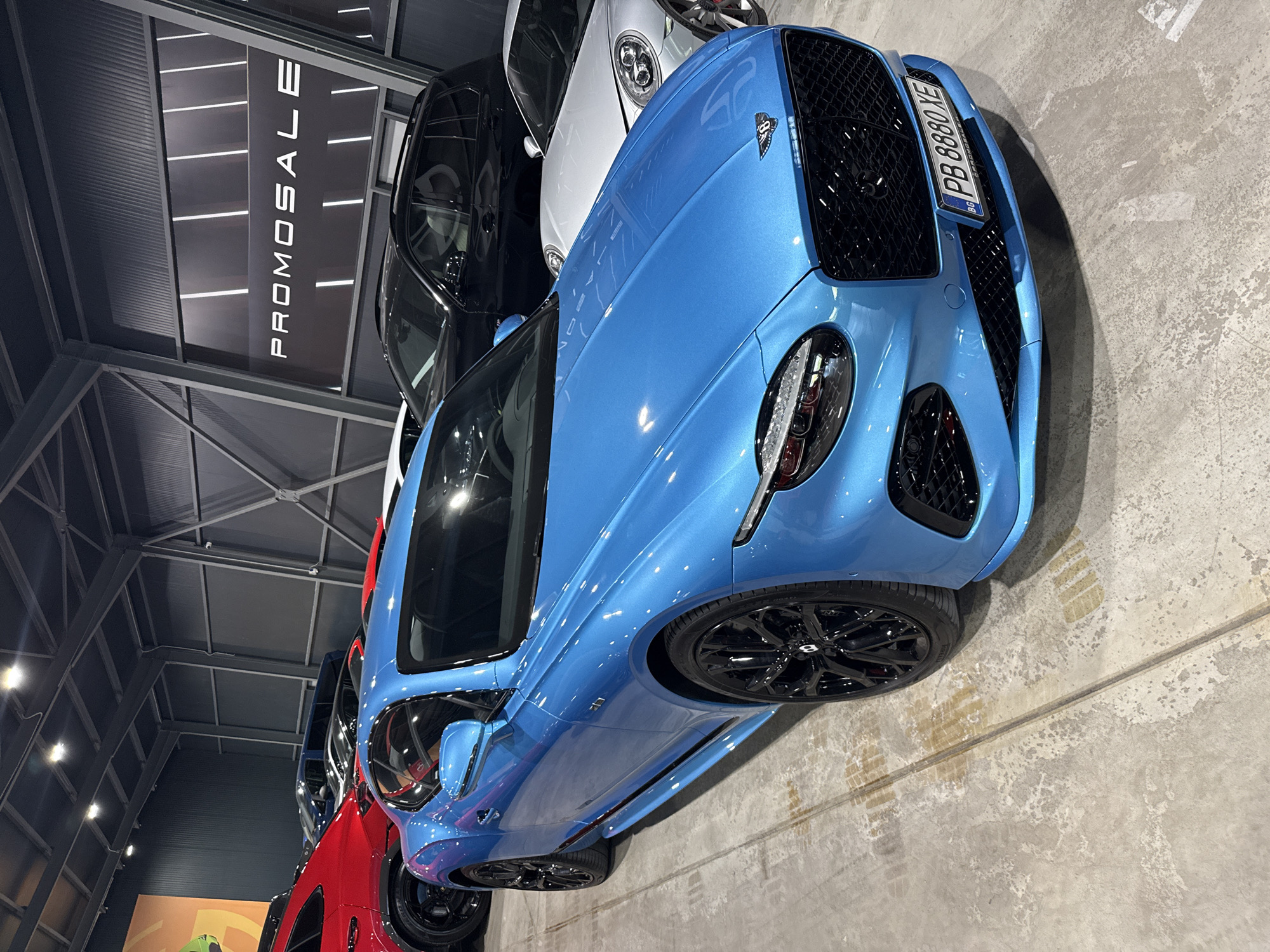 A bright blue Bentley Continental GT is parked on a concrete floor in a showroom. The car is angled towards the viewer, with its front left wheel prominently displayed. The car has black wheels and a black grille. A license plate with the characters "PB 8880 XE" is visible on the front of the car. In the background, other cars, including a red SUV, a black SUV, and a silver sports car, are parked. The showroom has a high ceiling with exposed metal beams and industrial lighting. A large banner with the word "PROMOSALE" written in white lettering is displayed on the back wall. The floor shows tire marks and some water stains. The location is likely a car dealership or exhibition space in Plovdiv, Bulgaria, based on the license plate format. There are no people visible in the image.