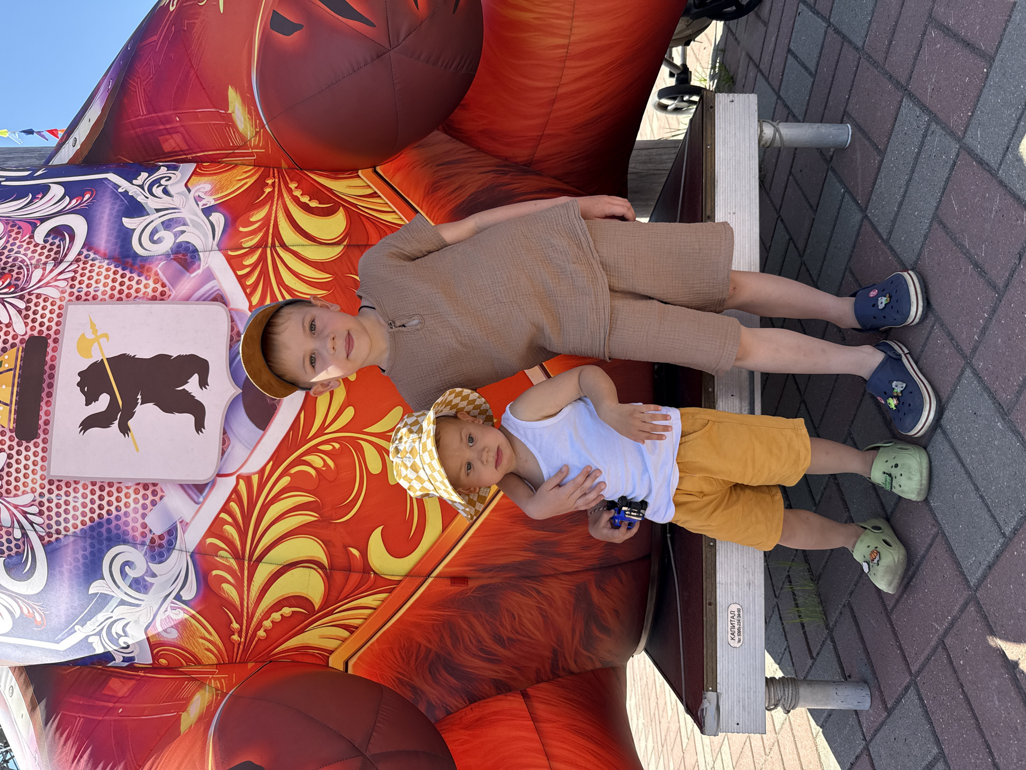 Two young boys, one older and one younger, stand side-by-side in front of a large, red and yellow inflatable structure featuring a bear emblem. The older boy, on the right, wears a tan t-shirt, tan shorts, and a tan baseball cap. He has his arm around the younger boy. The younger boy, on the left, wears a white tank top, yellow shorts, and a yellow and white checkered bucket hat. He is holding a small blue toy. Both boys are wearing clogs on their feet. They are standing on a raised platform with metal legs, in front of a brick-paved walkway. The inflatable structure is colorful and decorated with ornate patterns and the image of a bear holding a staff. The scene appears to be outdoors, under a clear blue sky. The text on the platform indicates the location is likely in Заволжье, Russia.