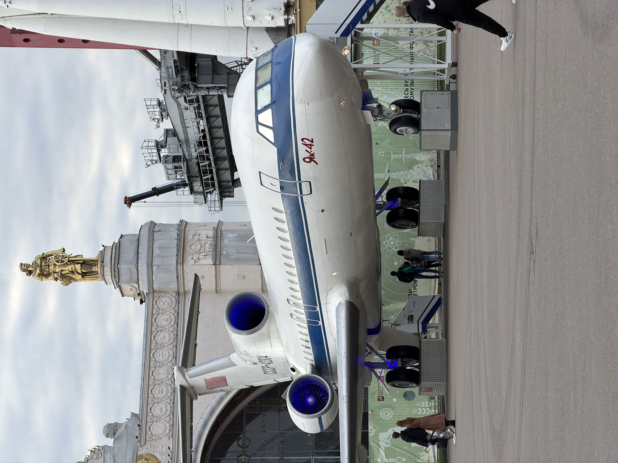 The image shows a Yak-42 airplane on display at the VDNKh (Exhibition of Achievements of National Economy) in Moscow, Russia. The plane is white with a blue stripe. The "Yak-42" is written on the nose of the plane in Russian. The building facade of VDNKh's Pavilion No. 1 "Central" and a rocket or rocket booster, perhaps related to space technology, are partially visible in the background. Several people are standing around the airplane, viewing it. The ground is paved, and there is a fence in the background, along with informational displays.