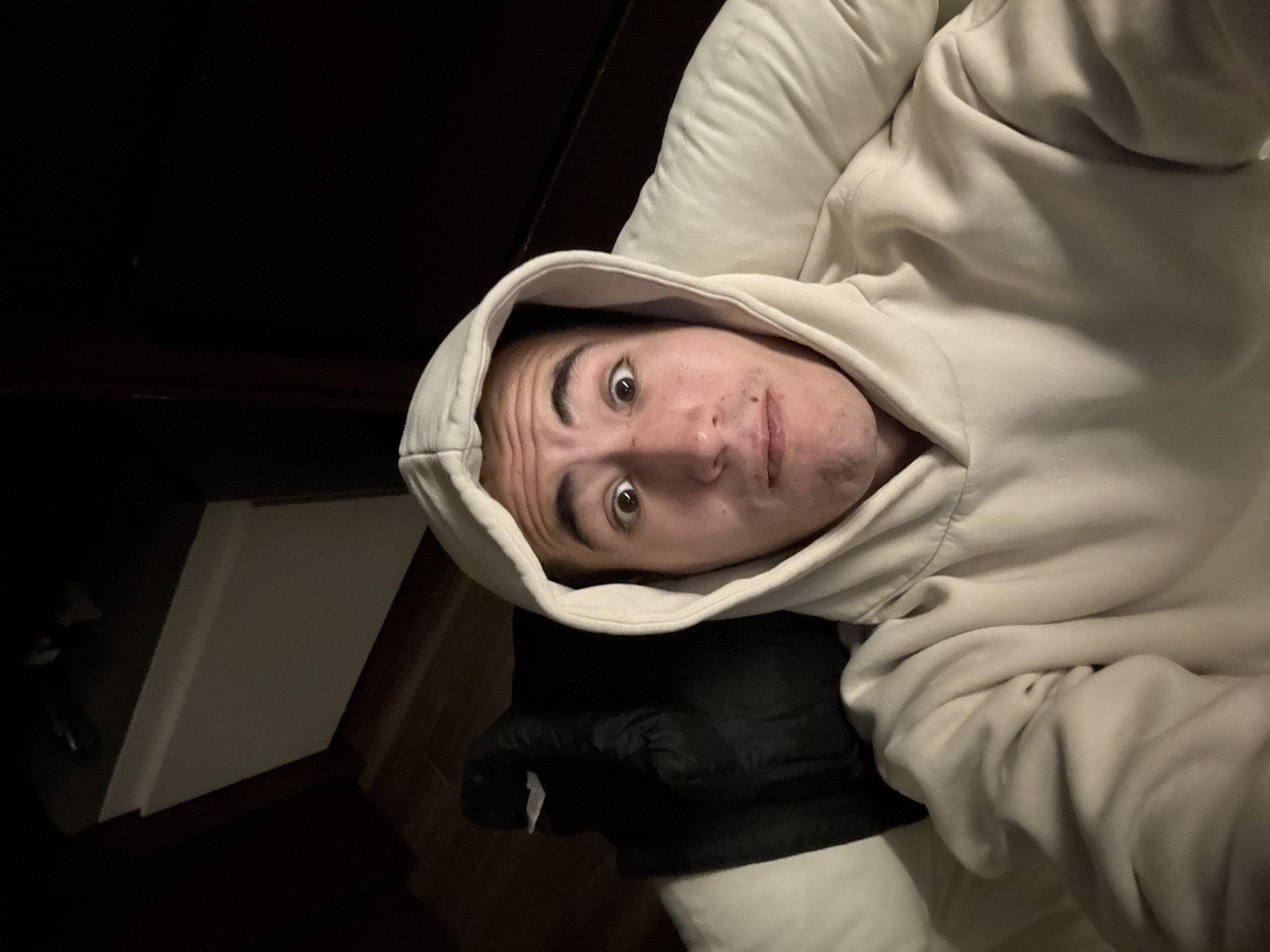 A young man is captured in a close-up shot, appearing to be lying down indoors. He is likely in his late teens or early twenties, with short dark hair, prominent eyebrows, and wide-open brown eyes that convey an expression of surprise, confusion, or a playful and curious gaze. His eyebrows are slightly furrowed, and his mouth is subtly parted.
He is wearing a light-colored, possibly cream or off-white, hooded sweatshirt, with the hood pulled up around his head. His head rests on a light-colored pillow or bedding, and a dark-colored, possibly black, blanket or coat is visible beneath him.
The setting is an indoor space, likely a bedroom, characterized by the bedding and the man's relaxed posture. The background is predominantly dark, suggesting either nighttime or a dimly lit room. Hints of dark brown furniture or wooden surfaces are visible in the top left and bottom right corners. On the floor or a surface in the bottom left, there is a white rectangular object, possibly a book or container.
The image captures a casual, intimate moment with low lighting. There is no discernible text within the image. The location context provided for this media is San Bernardino Tlaxcalancingo, Mexico.
