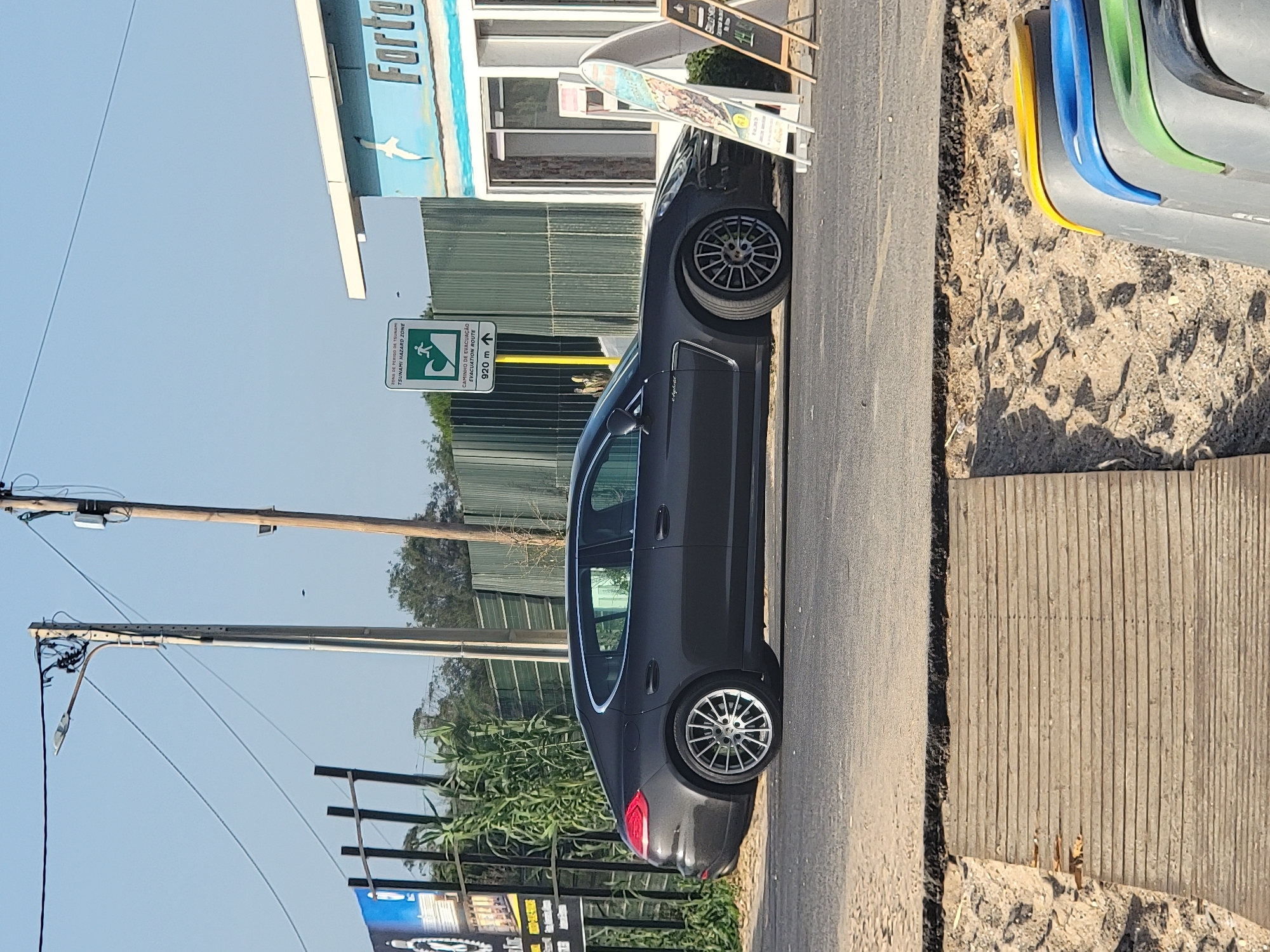 The image captures a dark gray Porsche Panamera sedan parked on an asphalt road in Quarteira, Portugal, near a beach. The car is positioned in profile view, facing towards the right of the frame. In the background, a blue and white building with the word "Forte" visible on it stands adjacent to a green metal fence. A tsunami hazard zone and evacuation route sign is prominently displayed to the left of the building. Several utility poles with electrical wires crisscross the sky. In the foreground, a wooden boardwalk leads towards sandy terrain where brightly colored recycling bins are visible. The scene appears to be outdoors during daylight.