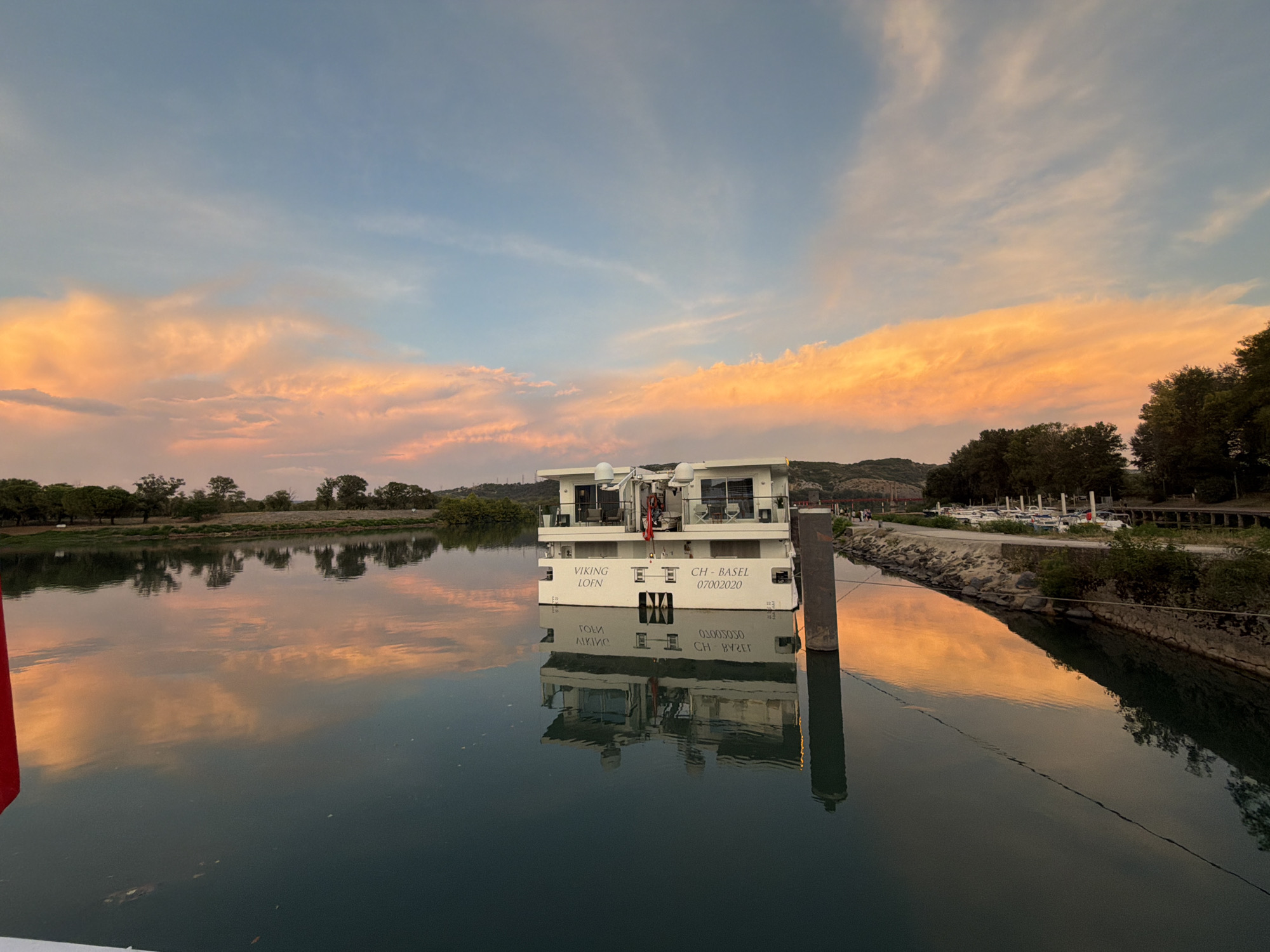 This is a wide-angle photograph of a river cruise ship docked on a calm river during sunset. The sky is filled with vibrant orange and pink clouds, reflected in the still water. The ship, a white vessel with multiple decks, is labeled "VIKING LOEN" and "CH-BASEL 07002020". A red flag is visible on the ship's deck. The riverbank is lined with trees and a stone embankment, with a road and parked boats visible in the background. The scene appears to be in or near the city of Viviers, France, likely on the Rhône River.