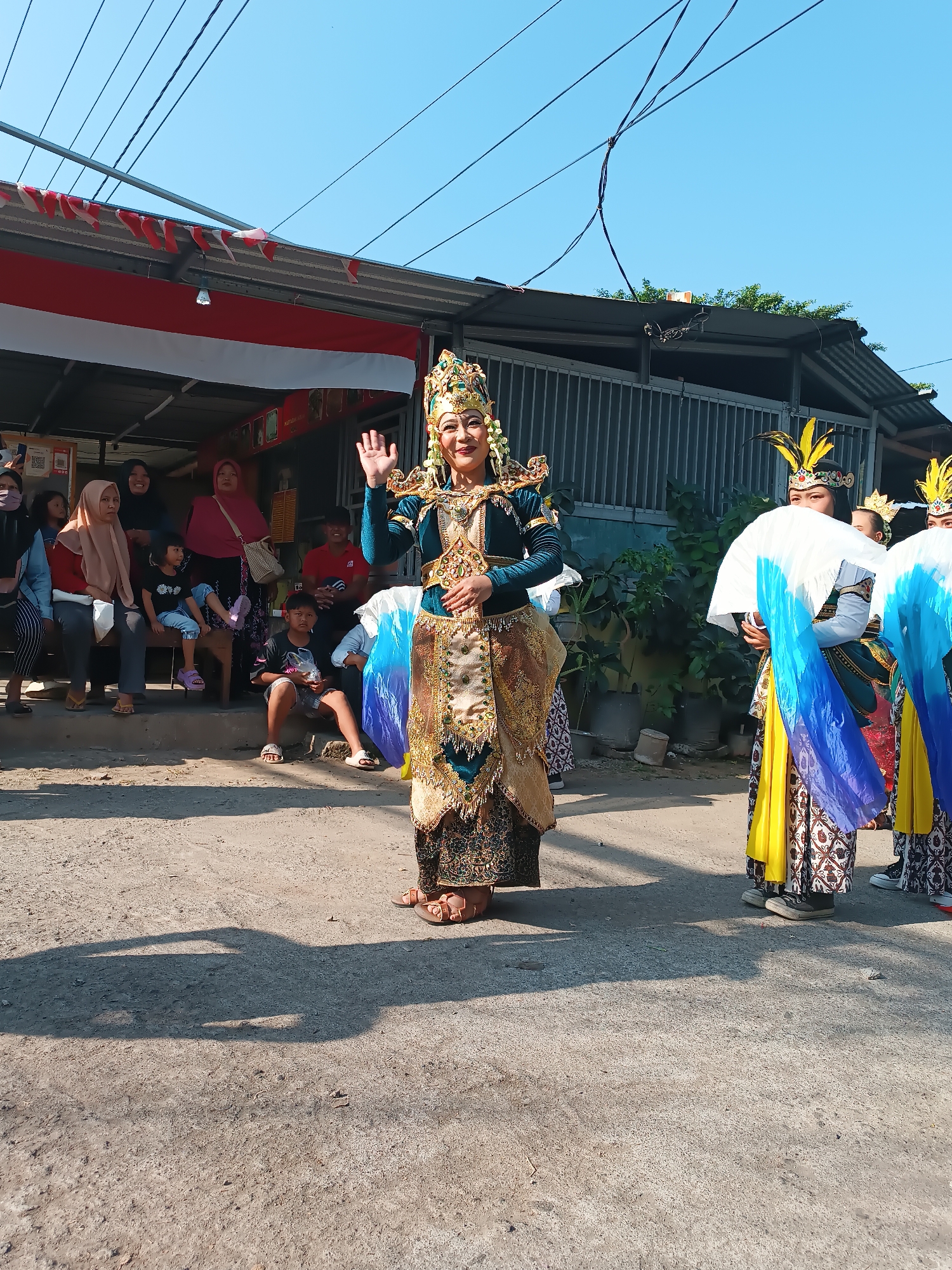 In Imogiri, Indonesia, a group of people are gathered on a street, likely for a cultural performance. The main subject is a woman in a traditional, ornate costume, including a tall, bejeweled headdress and a flowing, gold-embroidered outfit. She is standing in the foreground, waving to an unseen audience. Behind her, other performers in similar, though slightly different, traditional attire hold large, colorful fans. Spectators, including women in headscarves and children, are seated on a raised area to the left of the frame, observing the event. A long banner with red and white stripes, likely the Indonesian flag, is draped above the building in the background. The environment appears to be a typical street scene with a concrete surface and some greenery. The lighting suggests it is daytime.