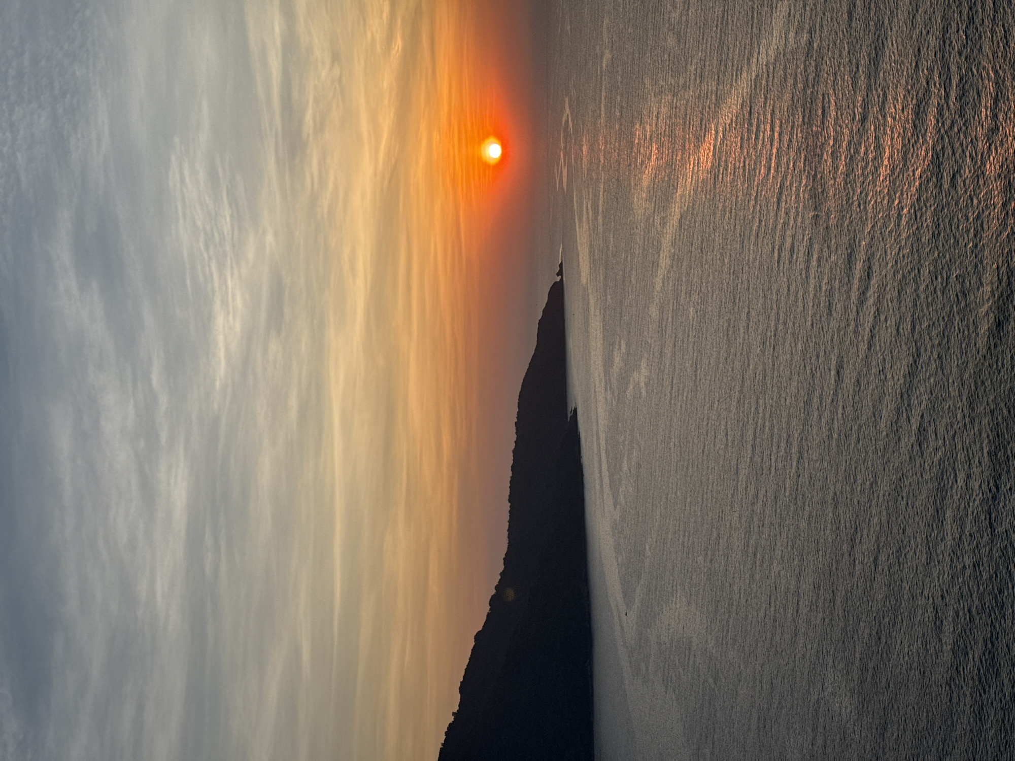 This image captures a serene sunset over a coastal landscape, likely viewed from a high vantage point overlooking the sea in Đà Nẵng, Vietnam. The horizon line diagonally separates the sky and the ocean.
The dominant feature is the bright, orange-red sun, positioned in the upper right quadrant, casting a warm and intense glow. The sky, occupying the upper portion of the frame, displays long, wispy clouds that transition in color from a soft grey-blue on the far left to vibrant yellows and oranges closer to the sun, indicating the late hour.
Below the sky, the expansive ocean stretches across the lower part of the image. The water's surface is textured with countless small ripples and waves, catching the light from the setting sun. A prominent path of bright, shimmering orange light, a reflection of the sun, extends across the water towards the left.
A dark, rugged mountain or landmass is silhouetted against the colorful sky, forming a striking profile along the lower middle and right side of the image. Its deep shadow contrasts sharply with the luminous sky and the sun's reflection on the water.
The time of day is clearly sunset, marked by the low position of the sun and the warm, golden hues in the sky and on the water. The weather appears clear but with some atmospheric haze or light cloud cover enhancing the colors. There are no people, objects, or visible text in the image, allowing the natural beauty of the scene to be the sole focus.