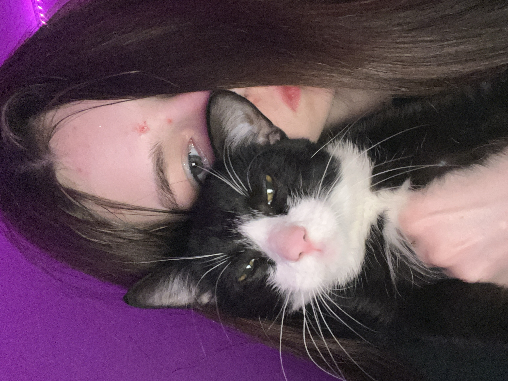A selfie-style image shows a person and a black and white cat. The person has long, dark brown hair and light skin. They have blemishes on their forehead and near their lip. The cat is lying on top of the person, with its face close to the camera. The cat has green eyes, a pink nose, and long white whiskers. The background is a blurry purple color. The image does not contain sufficient information to determine if the location is Saint Petersburg, Russia.