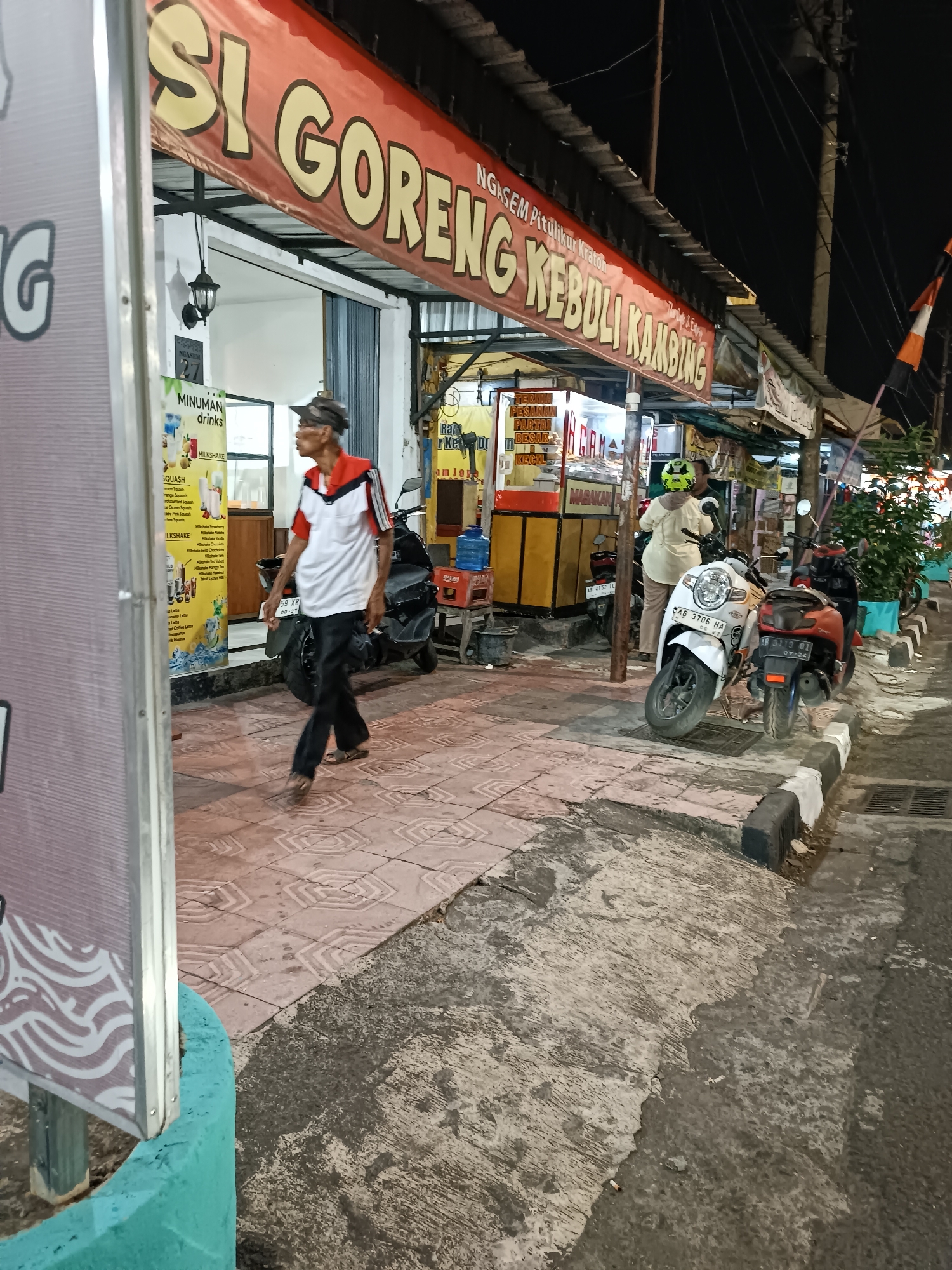 The image depicts a street scene at night in Yogyakarta, Indonesia. In the foreground, a man wearing a red and white shirt and black pants walks on a tiled sidewalk. A large red banner with yellow text, reading "SI GORENG KEBULI KAMBING" is prominently displayed above a food stall. Several motorcycles are parked along the sidewalk. One white motorcycle has a license plate that reads "AB 3706 HA". Another motorcycle, predominantly red, has a license plate "AB 3119 OI". Behind the motorcycles, a woman wearing a light-colored headscarf and a yellow top is interacting with someone at the stall. The stall itself has yellow and red signage and appears to be selling food. A menu board displaying "MINUMAN DRINKS" with various drink options is visible to the left of the stall. The environment suggests a commercial area with open-air food vendors. The ground is a mix of tiles and concrete, with some areas appearing wet. Electrical wires are visible overhead.