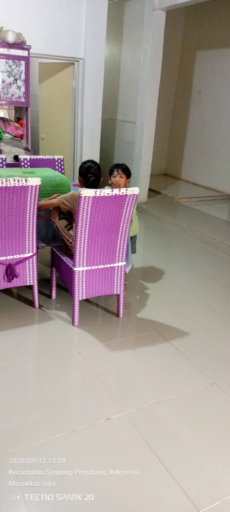 Two young children are seated at a table with purple woven chairs. The child on the right, facing the camera, has dark hair and is looking at the camera with a distressed expression, with hands near their mouth. The child on the left is turned away from the camera. A green cover is placed on the table. In the background, a tall, light-colored column is visible, along with a doorway. To the left of the children, a purple shelf unit with floral designs is partially visible. The scene appears to be indoors, with a tiled floor and shadows cast by the furniture. The timestamp and location indicate the image was taken on September 12, 2026, at 13:24 in Simpang Pematang, Indonesia.