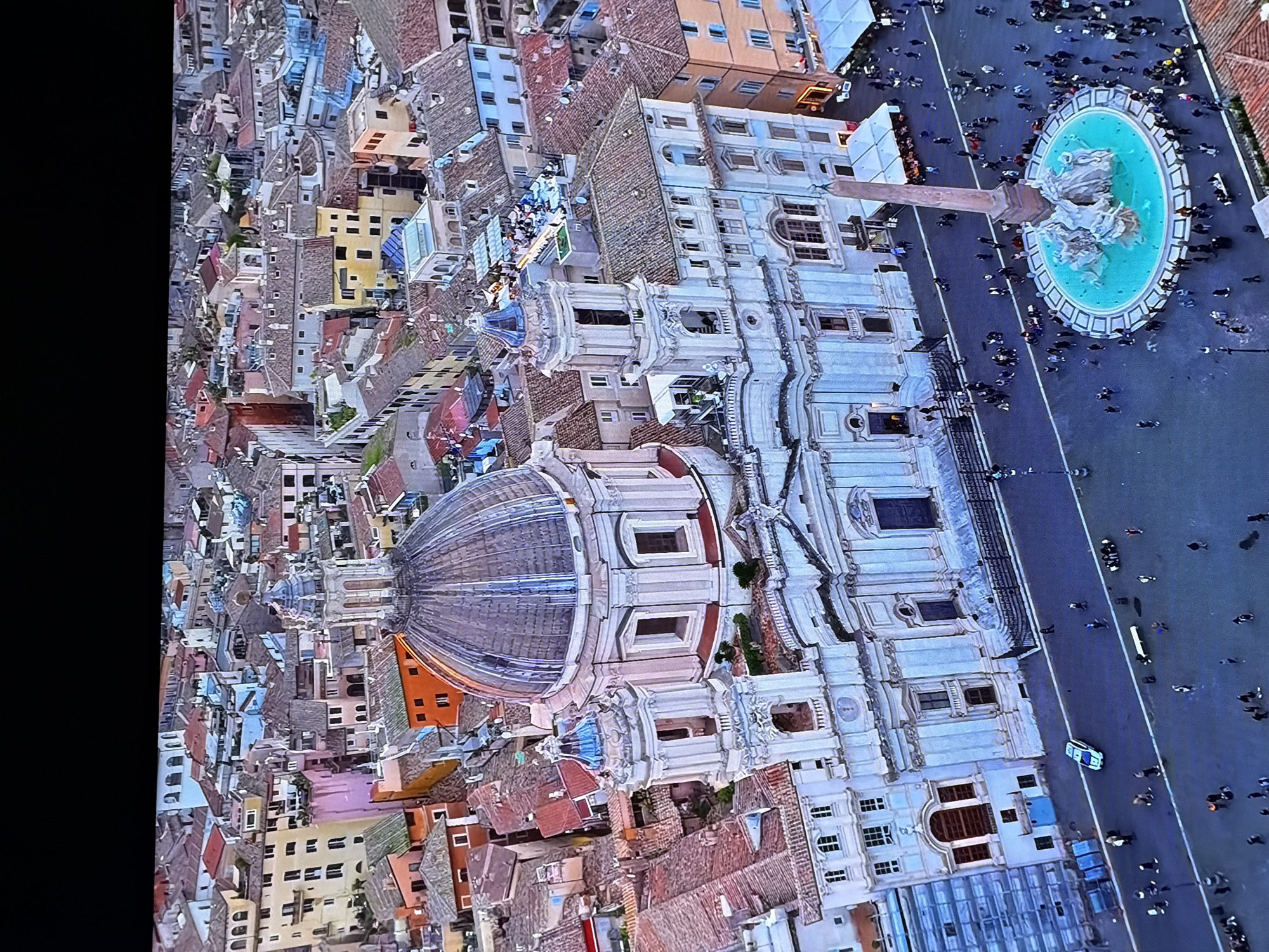 The image displays an aerial view of a city square, featuring a prominent Baroque church with a large dome and two bell towers. In front of the church, a large fountain with an obelisk at its center is surrounded by numerous people. The square is paved, and there are several buildings with terracotta roofs and light-colored facades surrounding the area. People are scattered throughout the square, some walking, some gathered, and some seated. The scene appears to be a public gathering space in an urban environment. The location is identified as the city of Chirchik, Uzbekistan.