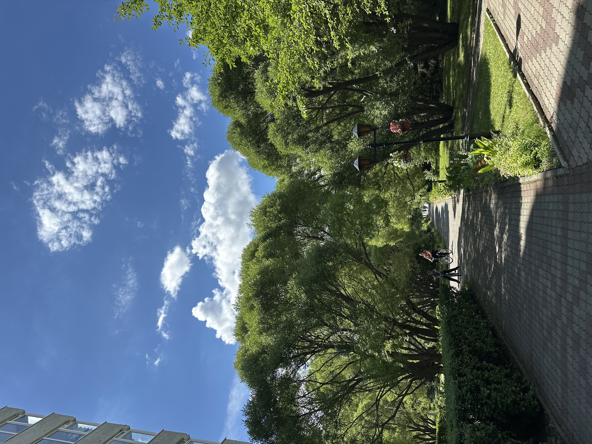 The image shows a paved pathway in a park in Duvankul', Russia, bordered by manicured hedges on the left and a grassy area with trees on the right. Two people are visible on bicycles on the pathway in the middle ground, and a building is partially visible on the far left. The sky above is blue with scattered white clouds. The pathway is made of red and grey paving stones. There are two lampposts with hanging flower baskets on the right side of the pathway. The trees are lush and green, indicating it is likely springtime or summer.