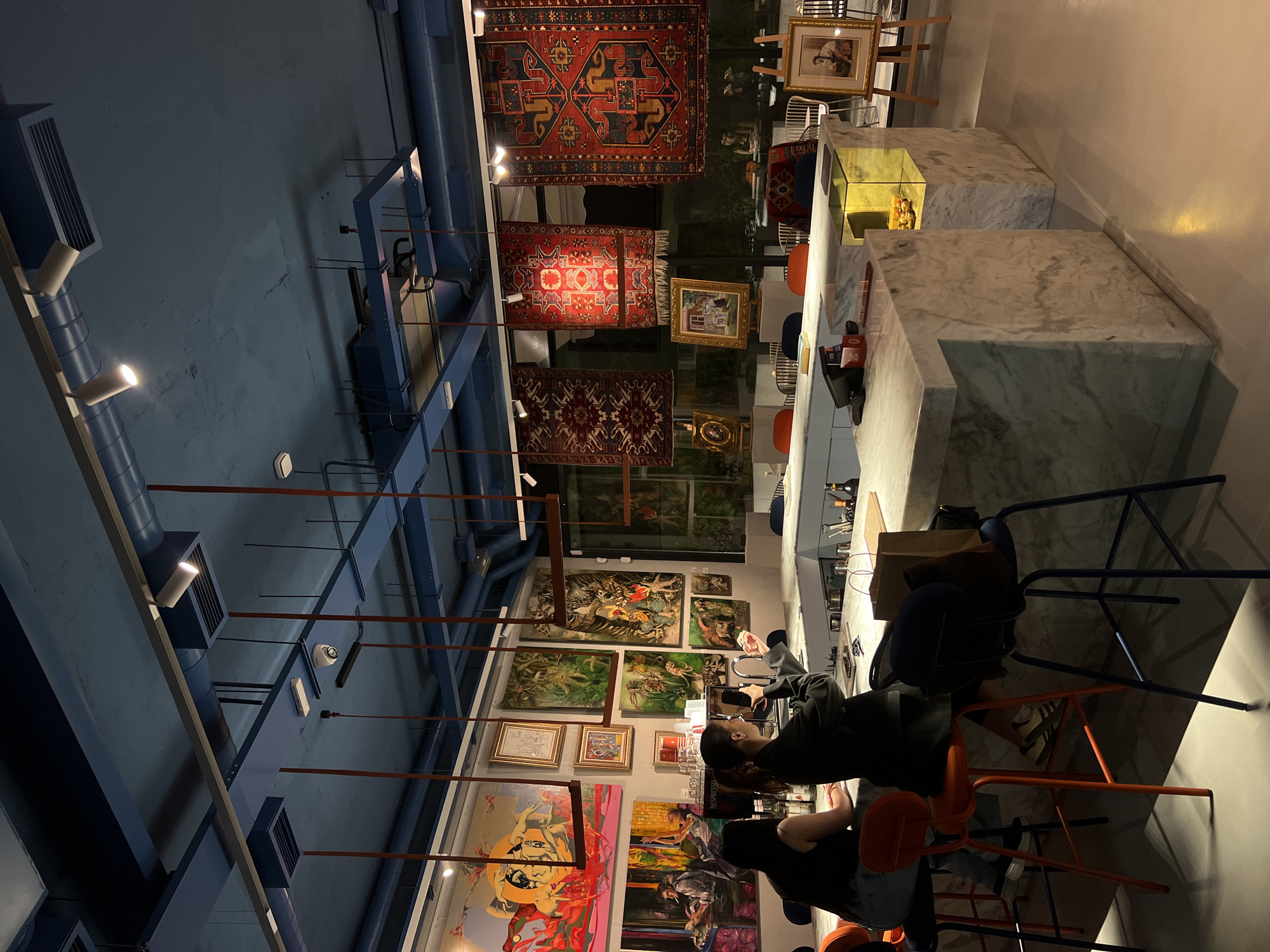 The image depicts the interior of a distinctive art gallery, cafe, or bar, likely in Yerevan, Armenia. The scene takes place indoors, appearing to be in the evening given the reliance on artificial lighting and the dark outdoor view through the windows.
The primary subjects include several individuals seated at a long, L-shaped white marble counter. One person, seen from the back, wears a dark jacket and sits on an orange stool, looking towards the wall adorned with art. Another individual is partially visible nearby, also seated at the counter.
The setting is characterized by a blend of artistic and industrial elements. The walls are densely covered with numerous framed paintings of various sizes and styles, showcasing a range of artistic expressions from abstract to illustrative. Overhead, several intricately patterned traditional carpets, likely Persian or Armenian, are hung from the ceiling, adding rich textures and colors. The ceiling itself reveals an industrial design with exposed dark blue pipes, air vents, and structural beams, complemented by track lighting fixtures that illuminate the space.
On the marble counter, various items are visible, including cups, bottles (one with "NESPRESSO" visible), a point-of-sale system, and a brown paper bag. A small, internally lit display case integrated into the counter holds an object. Seating consists of several bar stools, some in vibrant orange and others in dark blue or black.
The activity appears to be casual dining, drinking, or socializing. The ambiance is relaxed and sophisticated, encouraging patrons to enjoy the artistic surroundings. The overall impression is that of a modern, stylish venue that doubles as an exhibition space for art and traditional crafts.