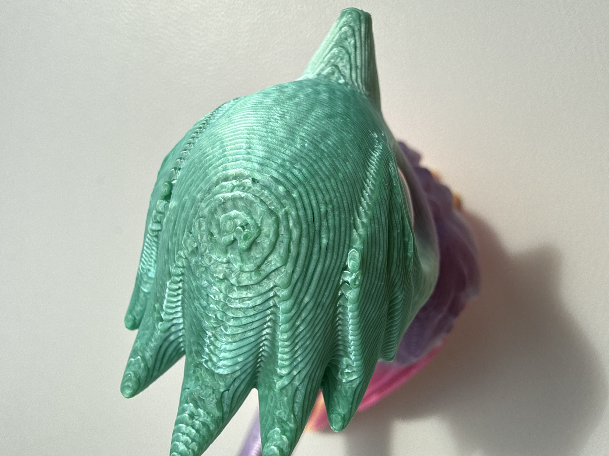 A close-up, top-down shot shows a 3D-printed object with a teal, textured surface. The object appears to be a stylized hand with visible layers from the printing process. It is positioned against a plain, light-colored background, with a soft shadow cast to the lower left. The bottom portion of the object shows glimpses of pink and purple colors, suggesting it is part of a larger figure. The scene is sterile and studio-like, with no indication of an urban environment or a specific city.