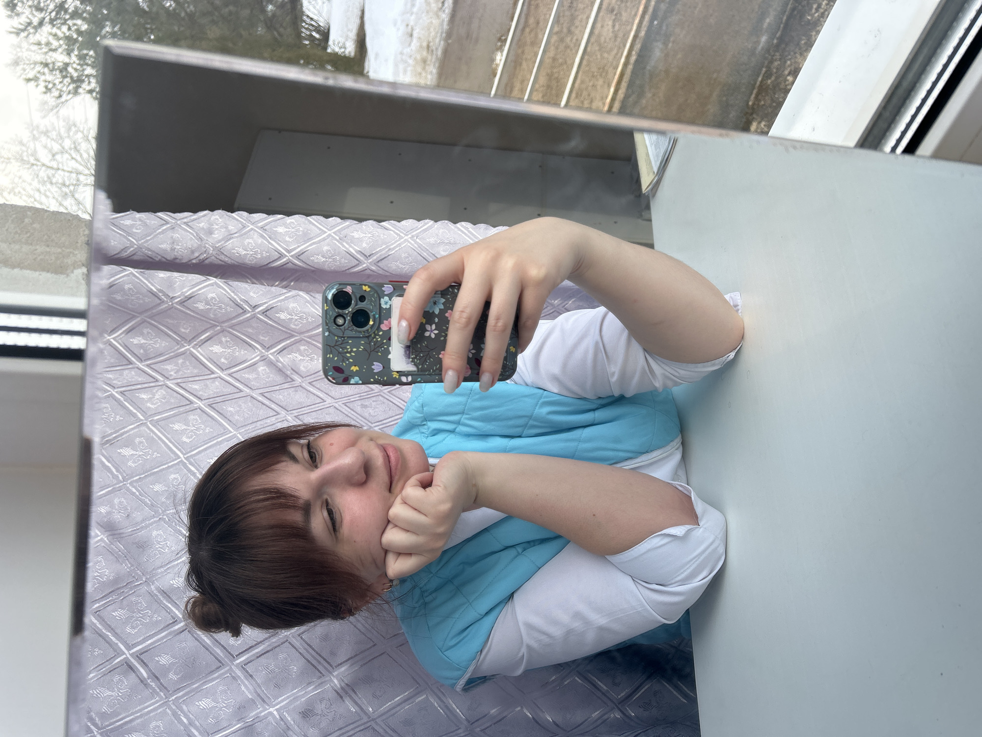 The image captures a female individual taking a mirror selfie indoors in the city of Фоки, Russia. The subject, visible from the chest up, has light-brown hair styled in a bun with bangs and wears a white long-sleeved shirt under a light-blue quilted vest. She holds a dark-colored smartphone, equipped with a floral-patterned case and three visible camera lenses, in her right hand, pointing it towards the reflective surface. Her left elbow rests on a white table, supporting her chin with her left hand. Her fingernails are manicured with white tips. Behind her, a light-colored curtain with a repeating diamond and fleur-de-lis pattern is visible. The reflection in the mirror also reveals an outdoor scene through a window, showing snow-covered ground and bare trees, consistent with a winter or early spring environment. The subject displays a subtle smile while looking at the smartphone screen.