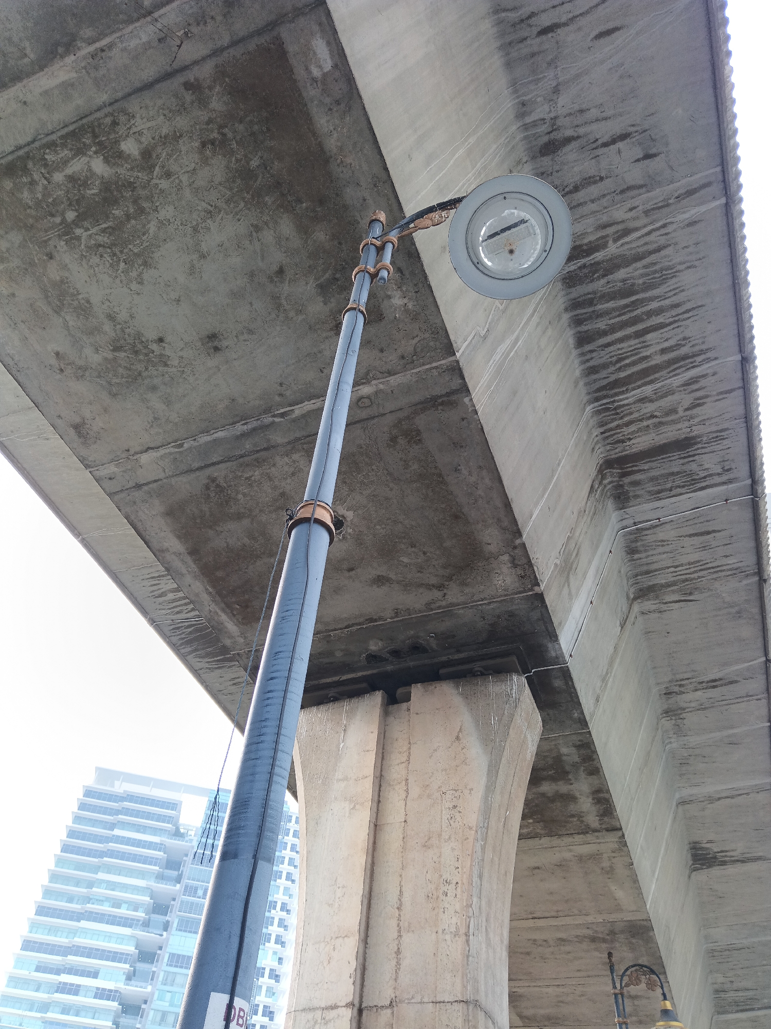 An upward-angle shot captures a utilitarian scene beneath a concrete overpass in Kuala Lumpur, Malaysia. A tall, dark gray lamppost extends upwards, its top adorned with a circular light fixture and a copper clamp. The lamppost is tethered by a blue rope. The overpass, a massive concrete structure, dominates the upper portion of the frame, showing signs of wear and tear with water stains and rough textures. To the left, a modern, multi-story building with glass windows and white balconies is partially visible. The lower right of the image reveals another, more ornate lamppost, partially obscured. The overall environment suggests an urban setting, likely a thoroughfare or public space within the city.