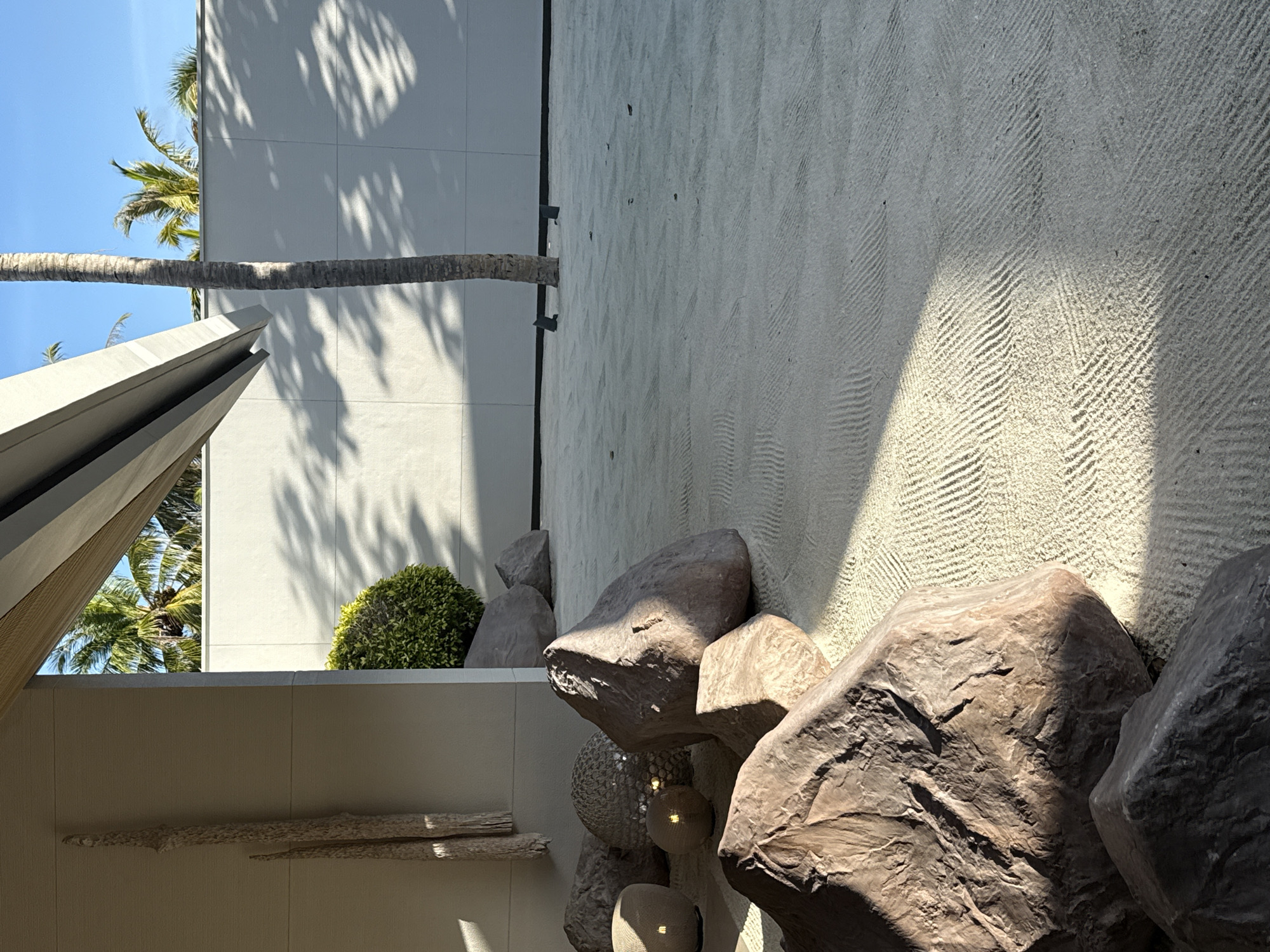 This is a medium shot photograph taken in a Zen garden. The scene is dominated by a smooth, light-colored wall with a textured pattern of diagonal lines etched into it, suggesting raked sand. A tall, slender palm tree trunk extends vertically through the frame, its shadow cast across the wall. To the left, there is a modern architectural element with sharp angles, possibly a wall or a roof overhang, also casting shadows. In the background, lush green palm fronds are visible, hinting at a tropical or subtropical location.
In the foreground, several large, rough-textured, dark brown rocks are arranged. Some of these rocks appear to be natural, while others might be sculpted or artificial. Beneath the rocks, there are decorative spherical objects, possibly lamps or sculptures, with visible light sources within them. A small, rounded green bush is positioned behind the rocks.
The lighting suggests it is daytime, likely morning or afternoon, with distinct shadows indicating sunlight. The overall mood is serene and contemplative, characteristic of a Zen garden. The presence of palm trees points towards a warm climate, and the mention of the Maldives as a potential location further supports this. There is no visible text in the image. The activity taking place is passive observation of the curated natural and architectural elements.