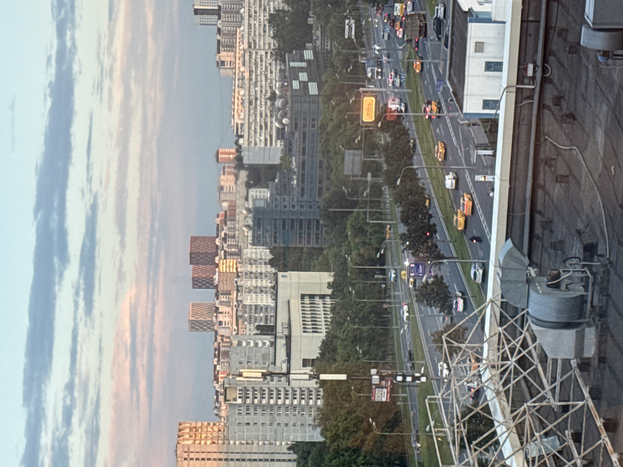 The image shows a view of a cityscape, likely Moscow, Russia, during what appears to be dusk. The foreground features the rooftop of a building with industrial ventilation equipment and a large metal structure. Beyond the rooftop, a multi-lane highway is visible with numerous cars, including yellow taxis, driving in both directions. Several buildings, varying in height and architectural style, are spread across the middle ground and background, some with visible apartment blocks and others appearing to be office or administrative buildings. Trees line the highway and surround some of the buildings, indicating a mix of urban and green spaces. The sky is partly cloudy, with streaks of pink and orange suggesting the sunset. Traffic lights and street lamps are also present along the highway. A few individuals are visible on the highway and near the buildings. The overall scene depicts a busy urban environment with ongoing traffic flow.