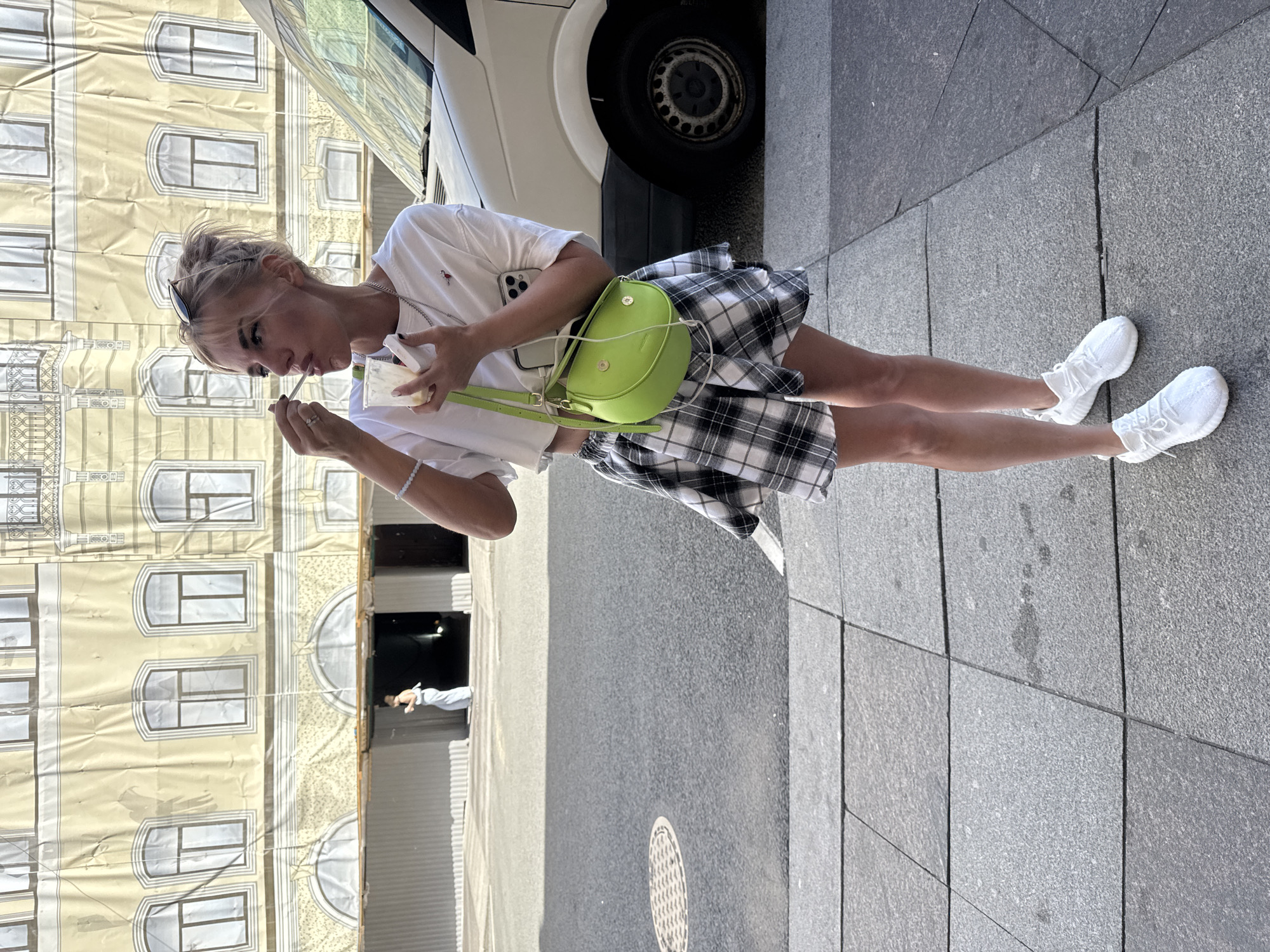 A young woman is standing on a sidewalk in front of a building with a facade depicting windows and architectural details. She is wearing a white t-shirt, a black and white plaid skirt, and white sneakers. She has a bright green crossbody bag. The woman is holding a small white object with a straw and appears to be drinking from it. She has blonde hair and is looking down at the object in her hands.
The building facade looks like it is covered by a large banner, possibly for construction or an event. To the left of the woman is the side of a white vehicle, possibly a van, with a visible tire. The sidewalk is paved with gray granite tiles. In the background, through an opening in the building's facade, another person can be seen. The scene appears to be outdoors, likely during the daytime, given the lighting. The overall impression is a casual street scene.