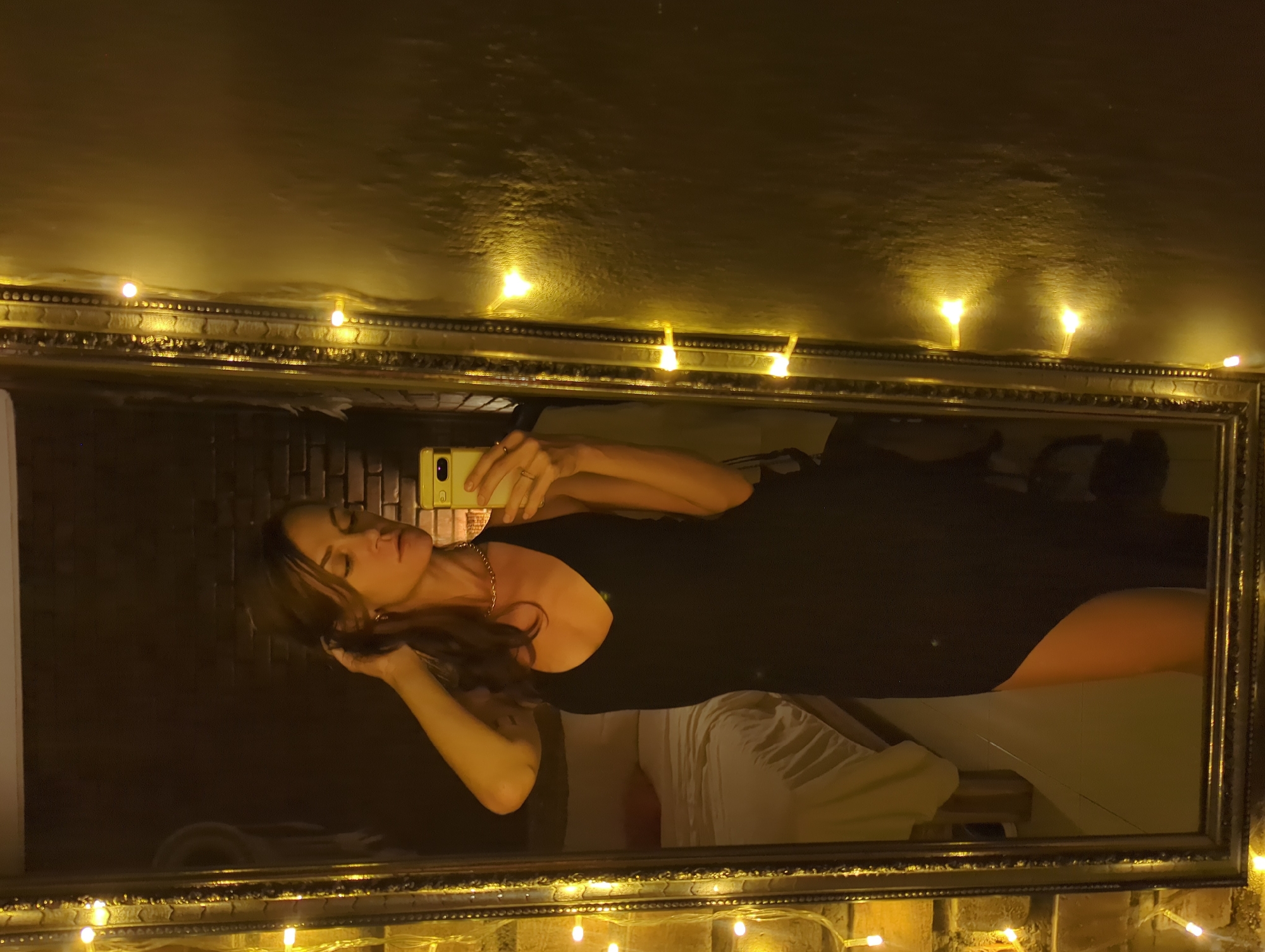 A woman is taking a selfie in a mirror, presumably in a private residence with exposed brick walls adorned with string lights. The mirror has an ornate frame. The woman is wearing a dark-colored outfit, possibly a dress or bodysuit, and a gold necklace. She is holding a smartphone in her right hand and her left hand is near her hair. A bed and white bedding can be seen in the reflection of the mirror. There is no confirmed evidence of this photo being taken in North Kuta, Indonesia.