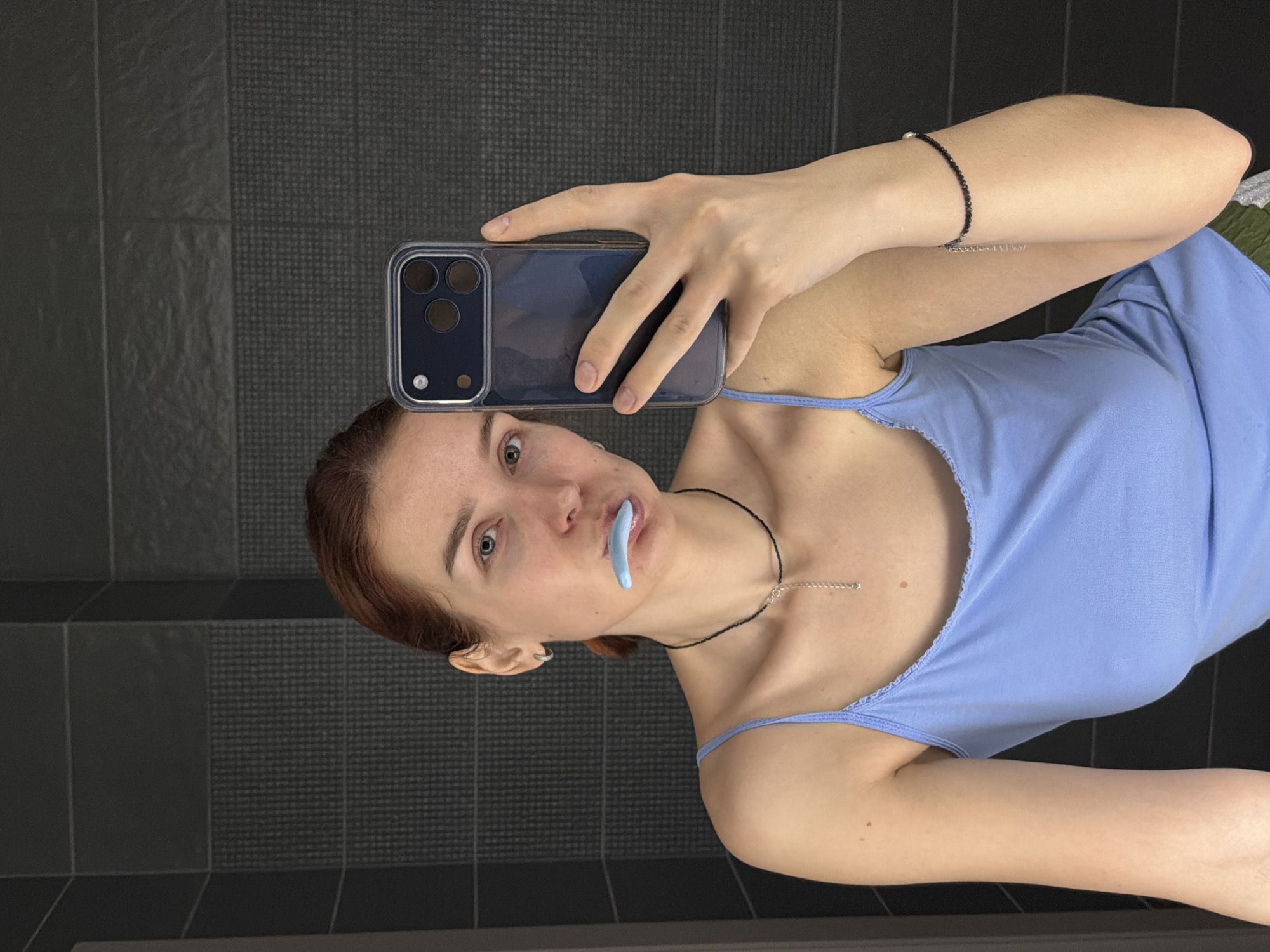 A woman is taking a selfie in a bathroom. She is wearing a light blue tank top and holding a smartphone with a blue case. She has a light blue dental device in her mouth. She has two necklaces on, one black and one silver. She has an earring in her ear and a bracelet on her wrist. She is standing in front of a gray tiled wall. The location is assumed to be in a bathroom of a residence in Chaikovsky, Russia.