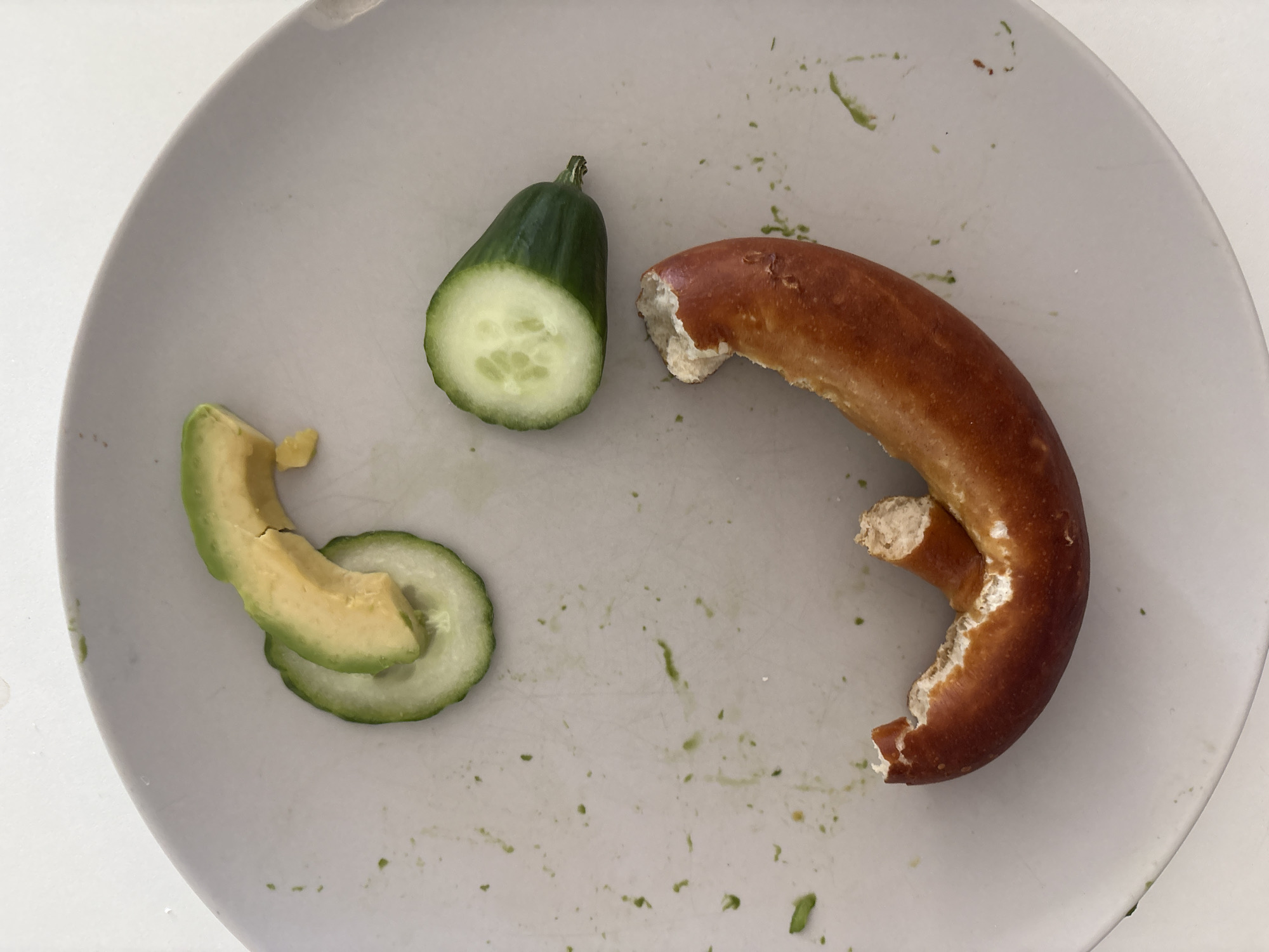 The image shows a light gray plate with several food items and remnants on it. The main subjects are a piece of cucumber, two slices of avocado, and a broken pretzel. The cucumber is a half-circle with a visible cut surface showing its seeds and white interior, with green skin. The avocado slices are light green and yellow, with a slightly creamy texture. The pretzel is a reddish-brown color with a chewy, doughy interior visible where it has been broken. There are small green flecks scattered across the plate, likely from the cucumber or avocado. The lighting suggests it is daytime. The overall scene depicts a partially eaten meal or snack on a plate. There is no visible text. The location context is Bönnigheim, Germany.