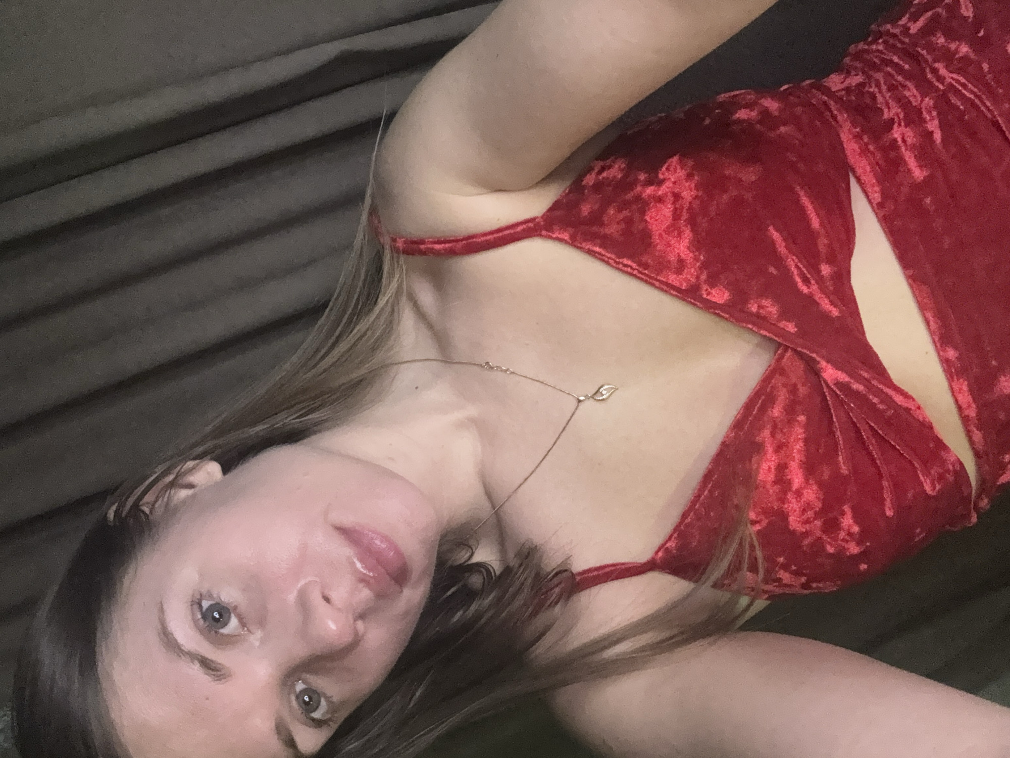 A light-skinned person with brown hair and blue eyes lies on a soft grey surface. They are wearing a red velvet dress with straps. A gold chain with a small leaf pendant is around their neck. The location is not identifiable as Batumi, Georgia.