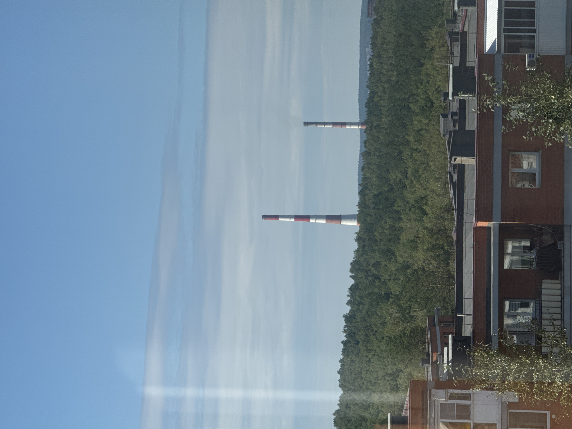 The image captures a daytime exterior view of an urban landscape, likely from an elevated position. The sky is a clear blue with sparse, wispy white clouds. Dominating the middle ground is a dense, green forest stretching across the horizon. Two tall, industrial chimneys, distinguished by their red and white striped patterns, rise above the treeline, indicating a potential industrial area or power plant. In the foreground, the rooftops of multi-story apartment buildings are visible. These buildings are constructed with dark brick and feature various rooftop structures such as ventilation shafts and possibly satellite dishes. The presence of apartment buildings suggests a residential area adjacent to the forested zone and industrial structures. The overall scene depicts a mix of natural and man-made elements within a Russian urban environment.