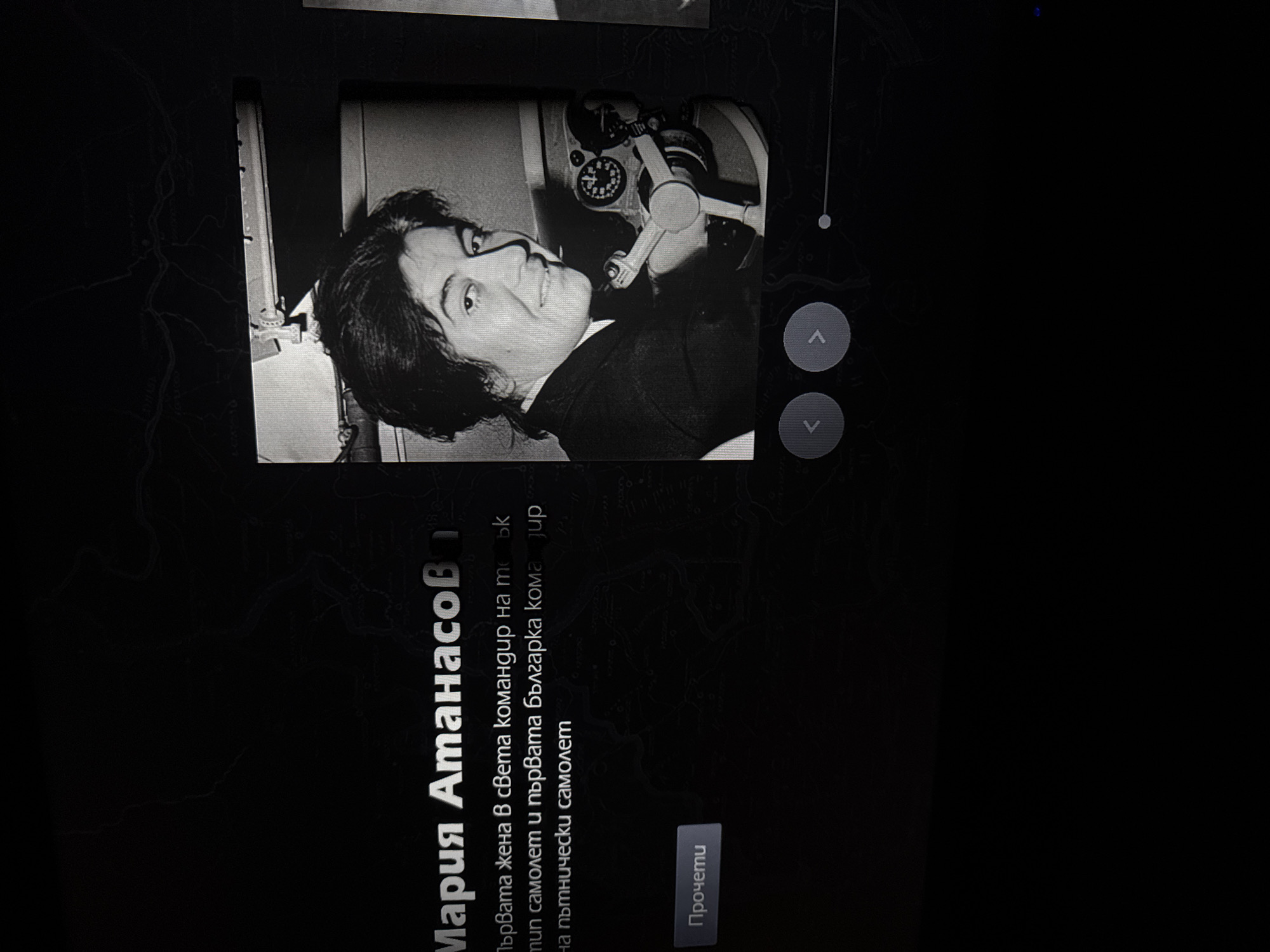 Here is a description of the image:
The image appears to be a screen capture showing an informational display. The focus is a black and white photograph of a person with dark hair, wearing a dark jacket or suit. They are situated next to what looks like an aviation instrument.
Adjacent to the photograph, there is text in what seems to be Bulgarian. The text includes the name "Мария Атанасов" (Maria Atanasov) and a description which translates to "The first woman in the world commander of (a specific) type of aircraft and the first Bulgarian commander of a passenger aircraft." There is also a button with "Прочети" (Read) on it.
The presence of the Bulgarian language indicates that the information pertains to something related to Bulgaria.
The photograph and text are centered on a background that appears to be a map or geographical illustration, although the details are obscured by the dark lighting and black overlay.