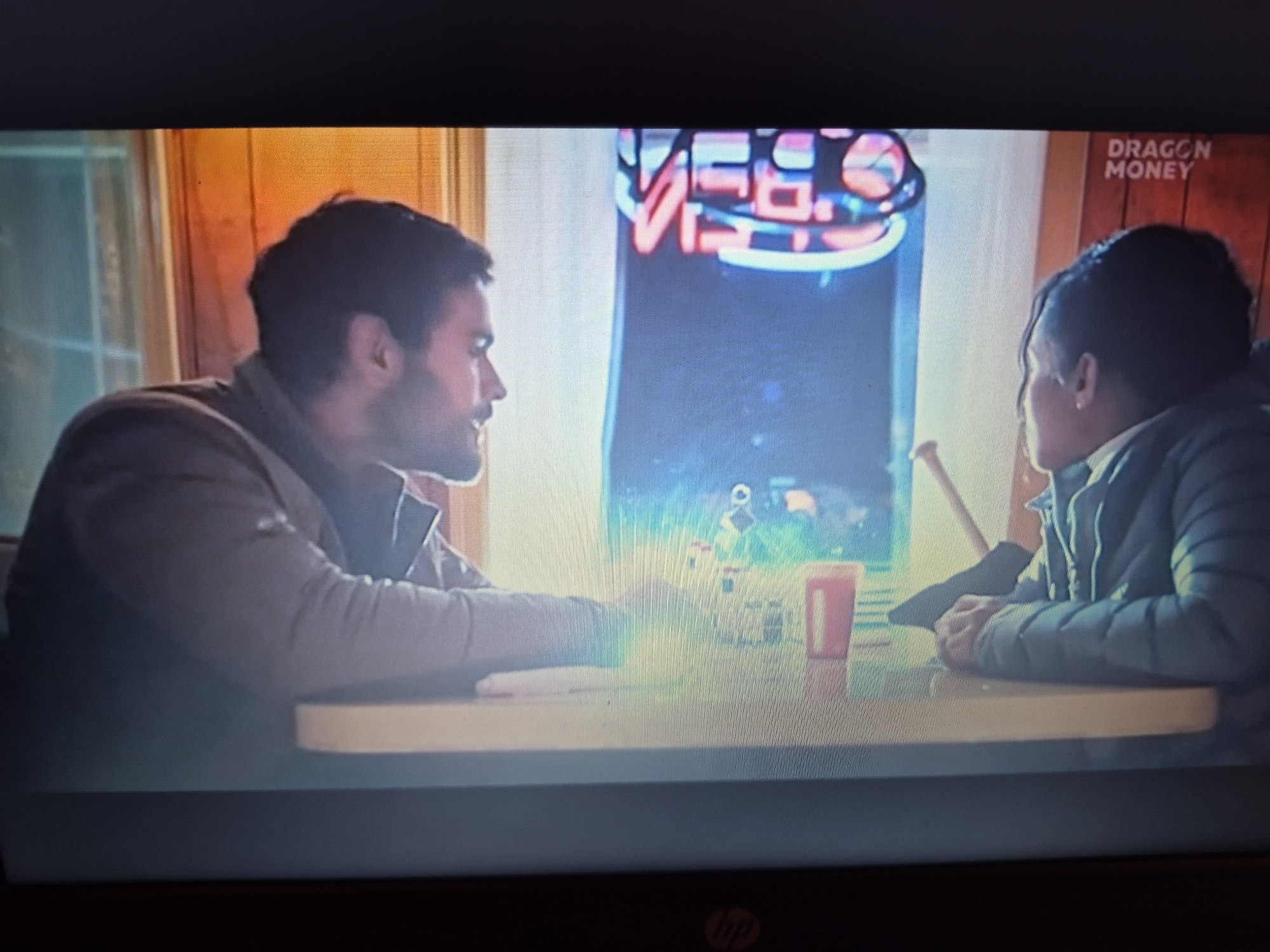 Two individuals are seated at a table in what appears to be a diner or lounge, illuminated by neon signage and warm-toned ambient lighting. The person on the left, a man with dark hair and a beard, is looking towards the right. He is wearing a dark jacket. The person on the right, with dark hair pulled back, is also looking towards the left. This person is wearing a dark, puffy jacket. Between them on the table are a red cup and what appears to be a white object, possibly a phone or a small book. Behind the individuals, a vibrant neon sign is visible, partially obscured by the person on the left. To the right of the neon sign, text in white lettering reads "DRAGON MONEY" against a wood-paneled background. The overall lighting suggests an indoor evening or nighttime setting. The location in the city of Sandwell, United Kingdom cannot be determined from the visual information provided.