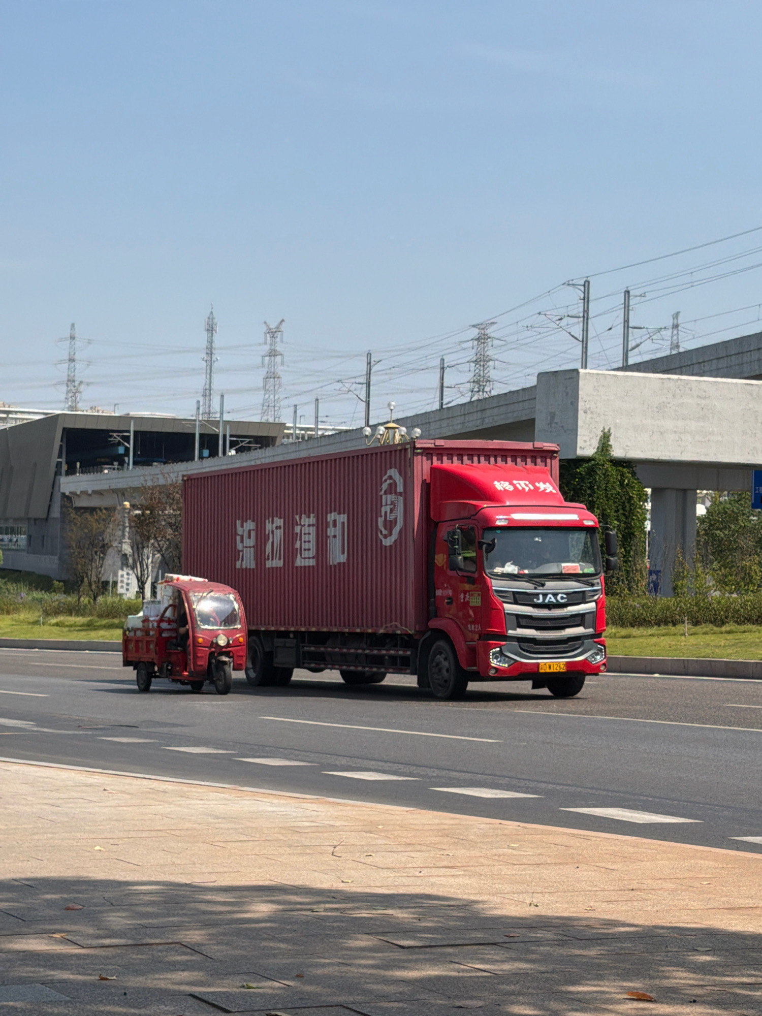A red, three-wheeled motorized rickshaw is driving on a road next to a red shipping container truck. The truck is a JAC model with the characters "渝运通达" on its side, which translates to "Yu Yunda". The license plate on the truck reads "川W1262". The rickshaw appears to be carrying some goods. In the background, there are power lines and a modern building structure, possibly a bridge or overpass. The sky is clear and blue. This scene appears to be located in the city of Jiangjin, China, given the "川" (Chuan) on the license plate, which is the abbreviation for Sichuan province, and the "W" which often indicates a city-level prefecture within Sichuan or Chongqing municipalities that border Sichuan. Jiangjin is a district of Chongqing, which was formerly part of Sichuan.