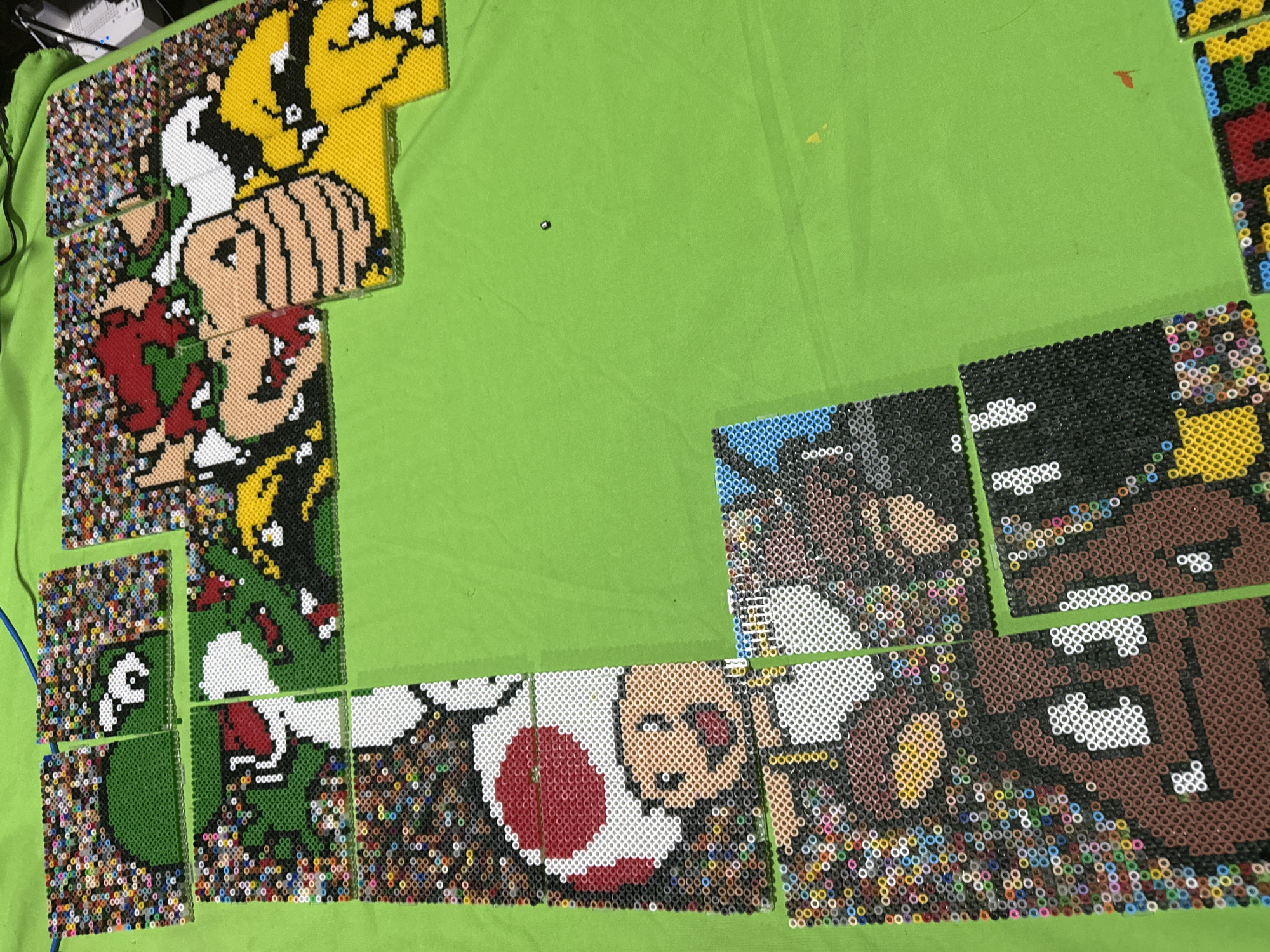 The image shows a partially completed perler bead creation featuring characters from the Super Mario franchise. The characters include Bowser, Mario, Yoshi, and Toad. The perler bead pieces are placed on a green fabric surface. The scene takes place indoors with overhead lighting visible.