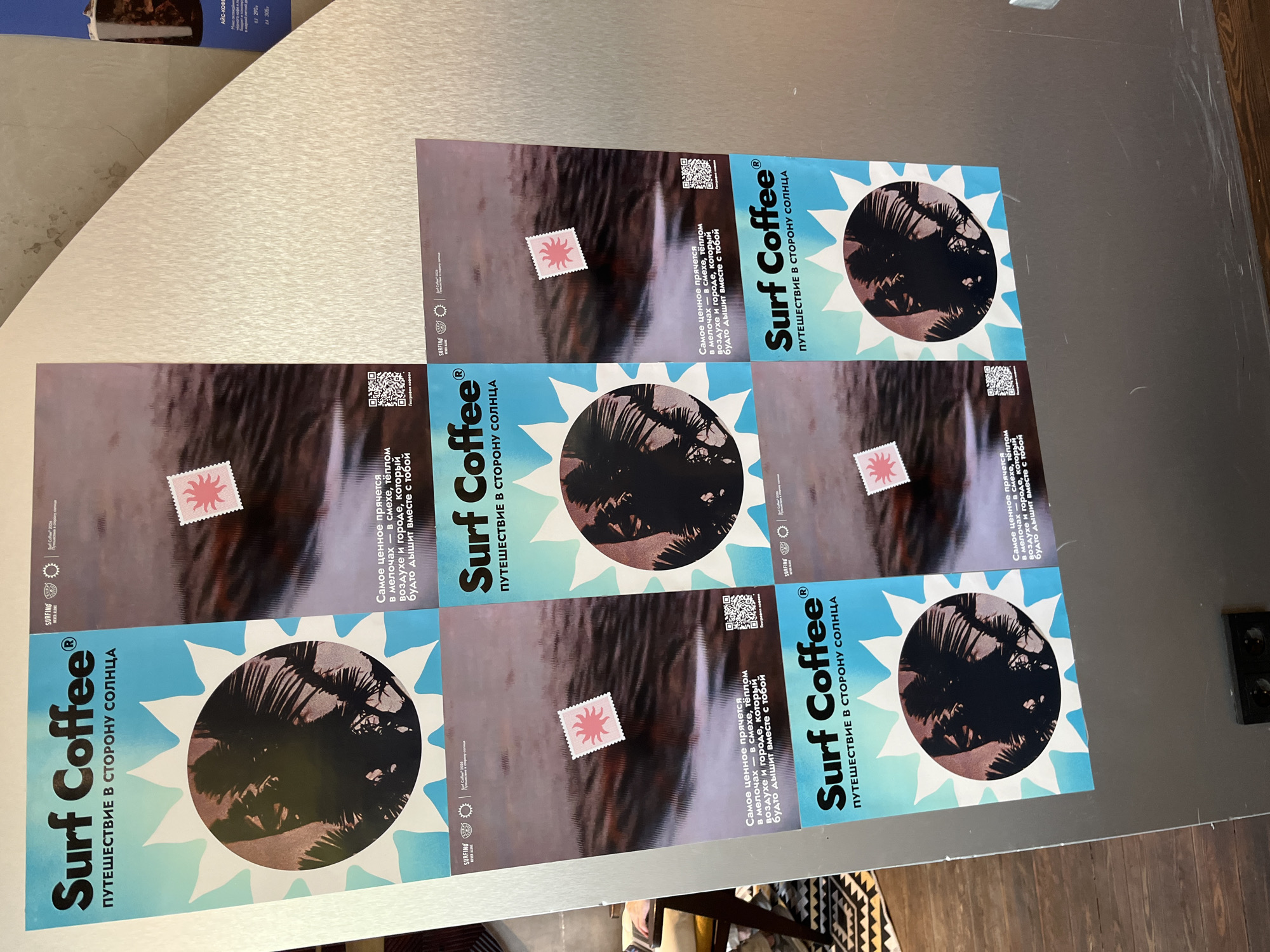 The image shows a wall with multiple posters for "Surf Coffee". The posters feature a sun graphic with silhouetted palm trees inside and the slogan "ПУТЕШЕСТВИЕ В СТОРОНУ СОЛНЦА" (Journey towards the sun). Some posters also include a small stamp-like graphic with a sun design, a QR code, and text. One poster in the upper right corner appears to be a menu or advertisement for "Surf Coffee" with details about coffee types and prices in Russian. The setting appears to be an indoor location, possibly a cafe or shop, with wooden flooring visible at the bottom of the image. The wall is light-colored and textured. The overall presentation suggests a promotional display for the coffee brand. The prompt mentions "city of Sochi, Russia" but there is no visual information within the image to confirm this location.