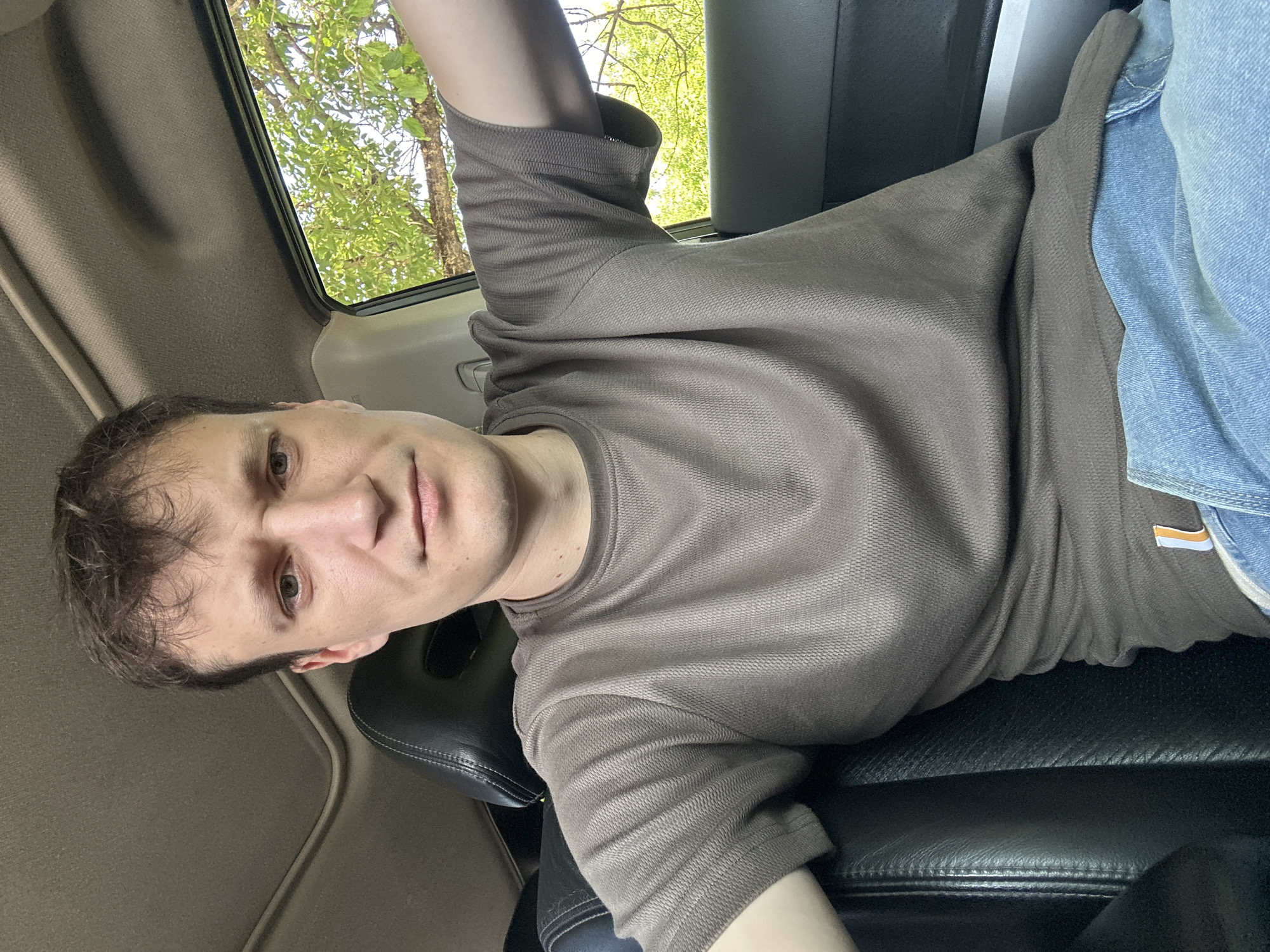 A Caucasian man with short, dark hair is sitting in the driver's seat of a car. He is wearing a short-sleeved, brown t-shirt and blue jeans. The interior of the car is visible, including the black leather seat and the car's headliner. Through the open car window, green trees and foliage are visible, suggesting an outdoor setting. The lighting indicates it is daytime, likely sunny. There is no specific information available to place this image in Makiv, Ukraine.