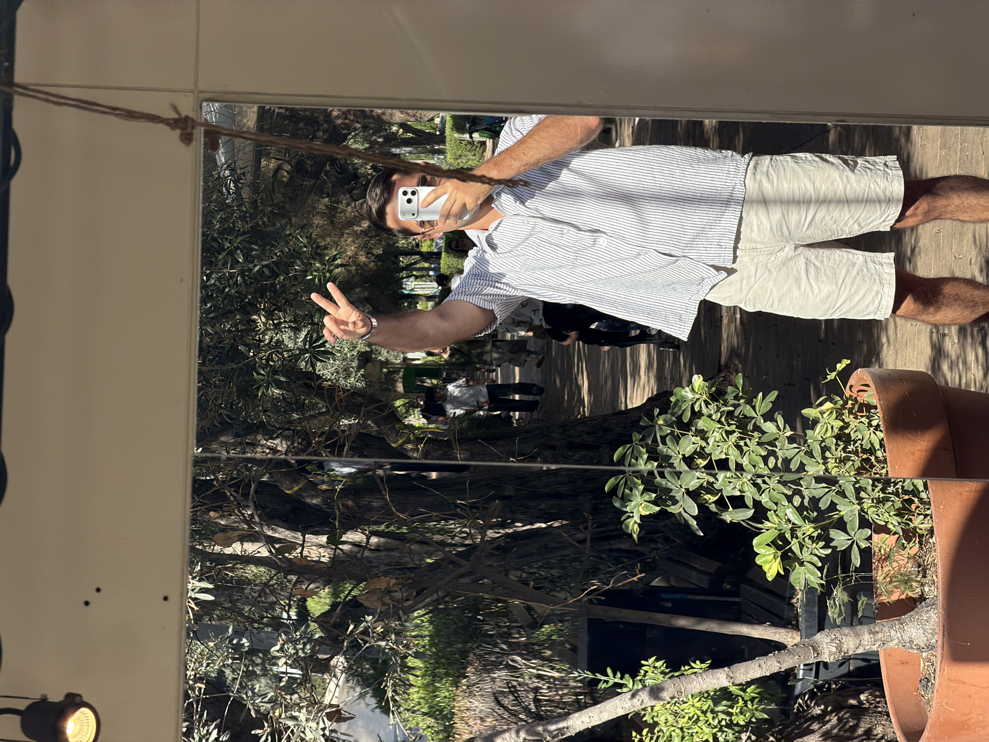 A man is captured in a mirror selfie, standing upright and making a peace sign with his left hand. He is wearing a short-sleeved, light-colored shirt with thin vertical stripes and light-colored shorts. A watch is visible on his left wrist. He holds a silver or white smartphone to his face to take the picture, partially obscuring his eyes.
The setting appears to be an outdoor or semi-outdoor area, reflected in the large mirror. Behind the man, the reflection shows a lush, green environment with numerous trees and various plants, suggesting a garden or patio. Other blurry figures of people are faintly visible in the background reflection, indicating a social gathering or a public space.
The ground where the man stands is a wooden deck or floor. To his lower right, a large terracotta pot with green foliage is visible. In the upper left portion of the mirror frame, a knotted rope or string is strung across. A small, illuminated spotlight is visible on the wall in the lower left corner.
The scene is brightly lit by natural sunlight, casting distinct shadows on the wooden floor, suggesting it is daytime, likely morning or afternoon. The weather appears clear and sunny. The man's pose and the overall atmosphere suggest a relaxed and cheerful mood. Given the location context, this scene is likely in Tel-Aviv, Israel, possibly in a garden, cafe, or private outdoor area. No discernible text is visible in the image.