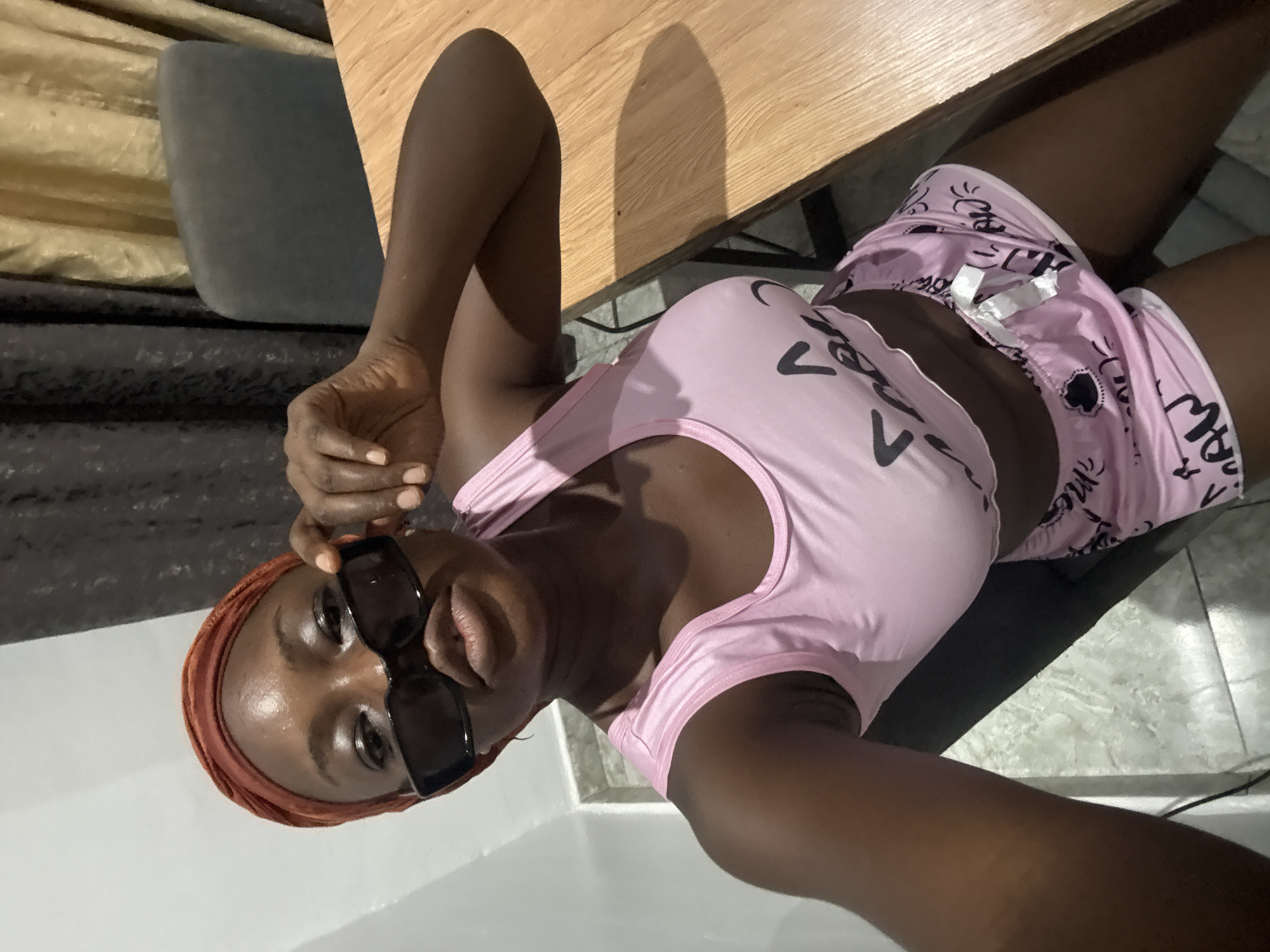A dark-skinned woman is depicted lying down, taking a selfie indoors in Iddo Tudunwada, Nigeria. She is wearing a light pink tank top and matching pink shorts adorned with black graphic patterns, an orange head wrap or scarf, and black sunglasses. Her head is slightly tilted, and she is looking directly into the camera with a neutral expression, her lips slightly pursed. One hand is visible near her head, suggesting she is holding the camera for the selfie.
She is positioned against a wooden surface, likely a table, which has a distinct shadow cast upon it. Behind her, elements of a room are visible, including a dark grey cushion or upholstered furniture, light-colored tiled flooring, and a pleated, golden-beige curtain or drapery in the background. The pink tank top features black graphic designs, including a prominent 'V' shape. The pink shorts are patterned with smaller black designs resembling hearts and stylized text or symbols, although specific words are not clearly decipherable. The indoor lighting suggests no specific time of day.