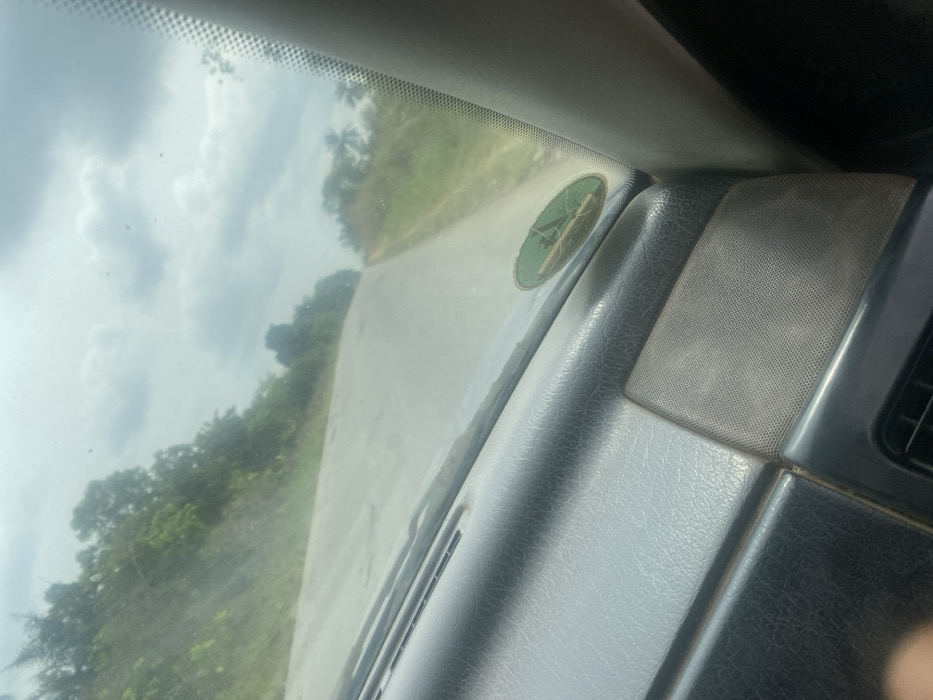 The image depicts a view from within a vehicle, looking forward through a windshield onto a rural road in Oka-Akoko, Nigeria. The vehicle's dark grey, textured dashboard occupies the lower foreground, displaying air vents and a speaker grille. A circular green decal with a white, possibly symbolic, design is affixed to the passenger side of the windshield, showing some wear. The windshield itself exhibits smudges and glare. Ahead, an unpaved or lightly paved light-grey road, showing surface imperfections, recedes into the distance, ascending a gentle incline and curving slightly to the right. Both sides of the road are bordered by dry grass and sparse vegetation, giving way to a dense expanse of green trees and shrubs, including palm trees on the right side. The sky above is overcast with scattered grey clouds. No individuals, overt actions, or interactions are visible.