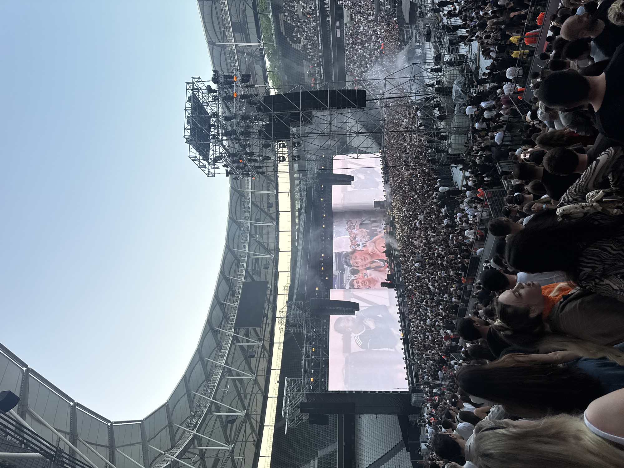 This image captures a large-scale outdoor concert within a modern stadium in an Unknown city. The sky above is clear and light blue, indicating late afternoon or early evening. The stadium features a distinctive curved, white canopy roof structure supported by an exposed metal truss system, and multiple tiers of grey seating.
The focal point is the concert stage at one end of the stadium. It includes a central performance area flanked by two very large rectangular LED screens displaying live video feeds. The left screen shows a male performer in a dark top, while the right screen depicts multiple individuals, possibly performers or engaged audience members. Two massive, multi-tiered metal truss structures, functioning as speaker and lighting towers, stand prominently in front of the stage, positioned further into the audience area. These towers are laden with numerous large speakers and various lighting fixtures. Smaller lighting rigs are visible above the main stage area. White smoke or haze effects emanate from the stage and near the speaker towers.
Thousands of people densely occupy the entire stadium floor in front of the stage, facing forward. The lower tiers of the stadium seating are also packed with spectators, while the upper tiers appear mostly empty. The audience consists predominantly of young to middle-aged adults, dressed casually in a mix of dark and neutral-colored attire. In the immediate foreground, several audience members are partially visible from behind or in profile, including a blonde-haired individual with a white strap top, a person with long dark hair, and individuals with an orange top and a patterned blazer, all observing the stage. Barriers separate sections of the crowd and stage infrastructure. Some utility personnel in high-visibility vests are visible near equipment on the stadium floor behind the main crowd.