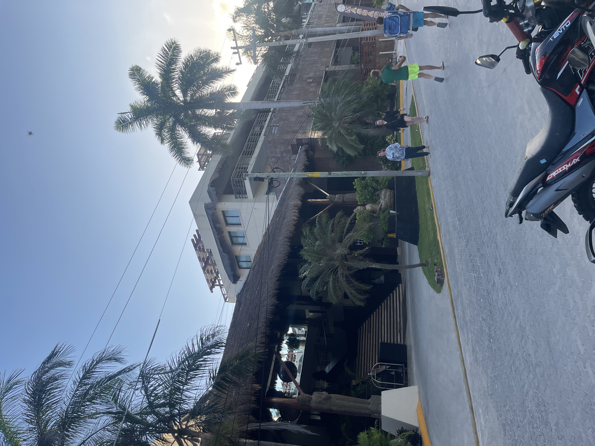 The photograph depicts a street scene in Playa del Carmen, Mexico. A three-story building with a thatched roof structure extending over a portion of the sidewalk. Several people are walking along the sidewalk, including a person in green shorts, a woman in black clothing, and two men farther up the street. A motorcycle is parked on the right side of the street. Palm trees are interspersed along the street and in front of the building. The sky is bright, suggesting it is daytime.