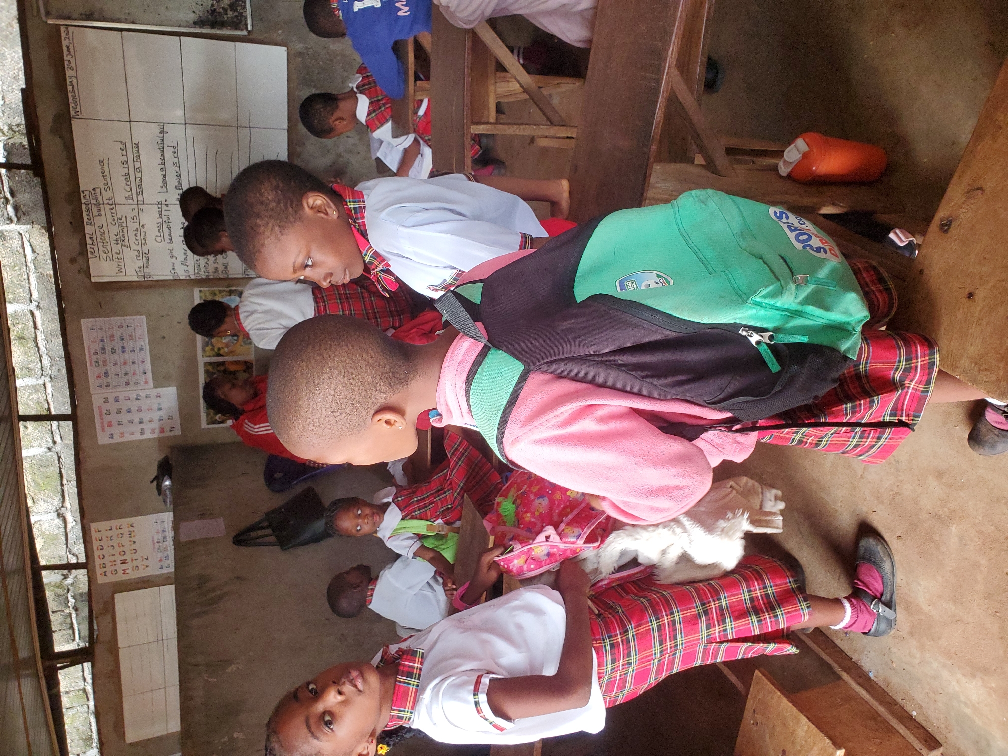This media file depicts a bustling classroom scene in Ukwu-Abwa, Nigeria, filled with young students engaged in learning. The setting appears to be a modest schoolroom with unpainted concrete block walls and simple wooden benches and desks. Natural light illuminates the room, suggesting it is daytime.
The primary subjects are numerous children, likely primary school-aged, dressed in matching school uniforms. The uniform consists of white collared shirts with short sleeves, paired with red and black plaid skirts or trousers, and plaid neckties or scarves. Many children are seen with their backs to the camera or in profile, appearing focused on their lessons. A boy in the foreground wears a vibrant green and black backpack. One child in the lower left foreground looks directly at the camera.
On the walls, two whiteboards are visible. The left whiteboard displays alphabet charts in both uppercase (ABCDEF, GHIJKL, etc.) and lowercase letters (Aa Bb Cc Dd, etc.), along with numbers (1234567). The larger whiteboard on the right has "Wednesday 3rd June, 2020" written at the top. Below this, it outlines a lesson on "Verbal Reasoning" and "Sentence building," providing instructions to "Write the correct sentence." Examples shown include:
- "The red crab is = The Crab is red"
- "I saw a house = I saw a house"
- "Class work"
- "Saw a beautiful girl = Saw a beautiful girl"
- "The flower is red."
Further down, there are blank lines and partial words like "house," "Friend," "bu s," and "Ball Ca" for additional exercises.
The children appear to be actively participating in the class, possibly writing or listening to instructions. An orange plastic container is visible on the floor in the background. The scene conveys an atmosphere of dedicated education in a basic but functional learning environment.