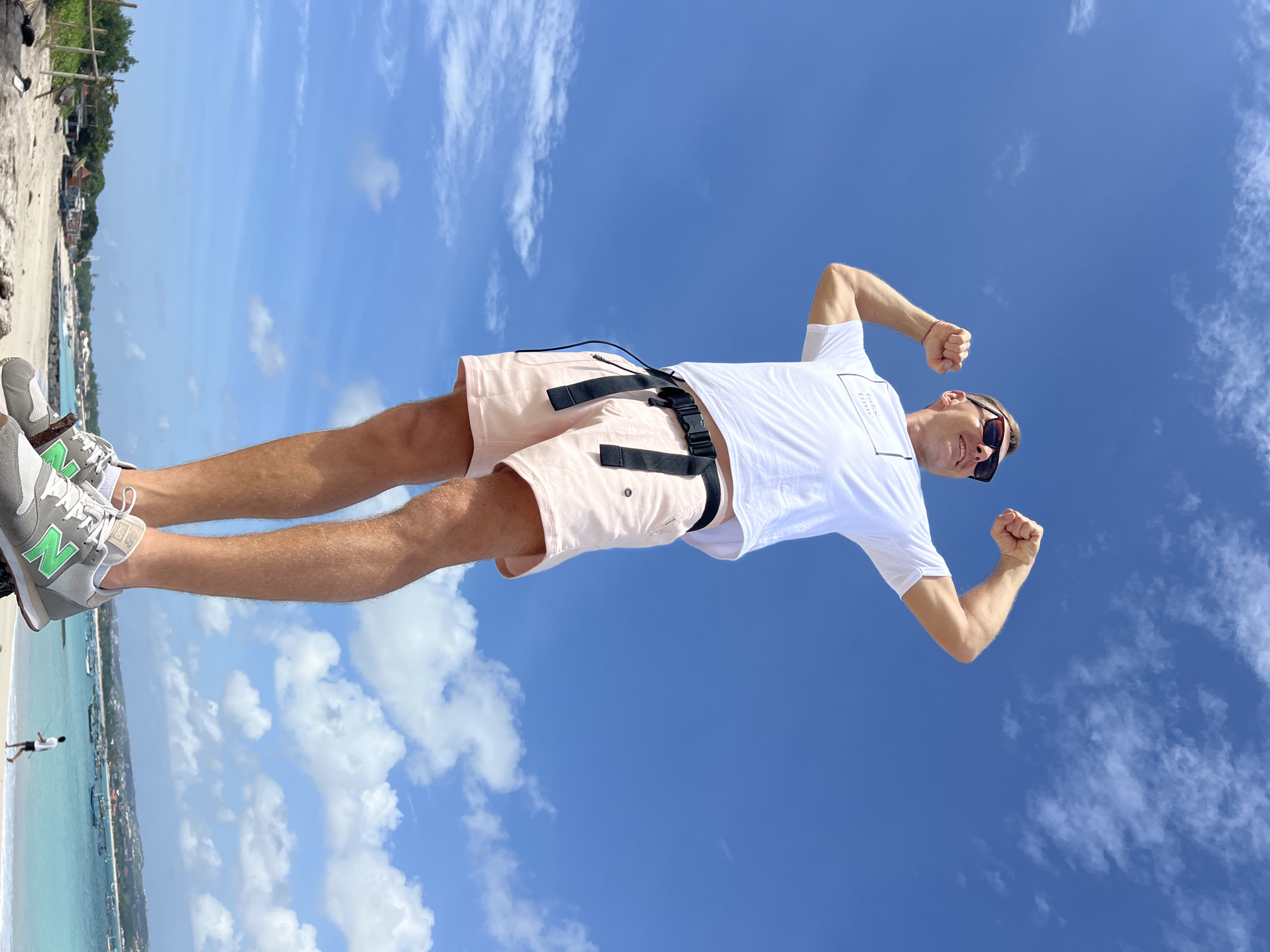 A full-shot, low-angle, eye-level outdoor photograph captures a man standing on a raised surface, looking up at the sky, and flexing his biceps. He is wearing sunglasses, a white t-shirt with a small graphic, light pink shorts with a black utility belt, and grey New Balance sneakers. The background features a clear blue sky with scattered white clouds, a beach, the ocean with several boats, and a distant coastline with buildings and vegetation. The man appears to be on a beach in Denpasar, Indonesia, given the visible tropical scenery and boat types commonly found in the region.