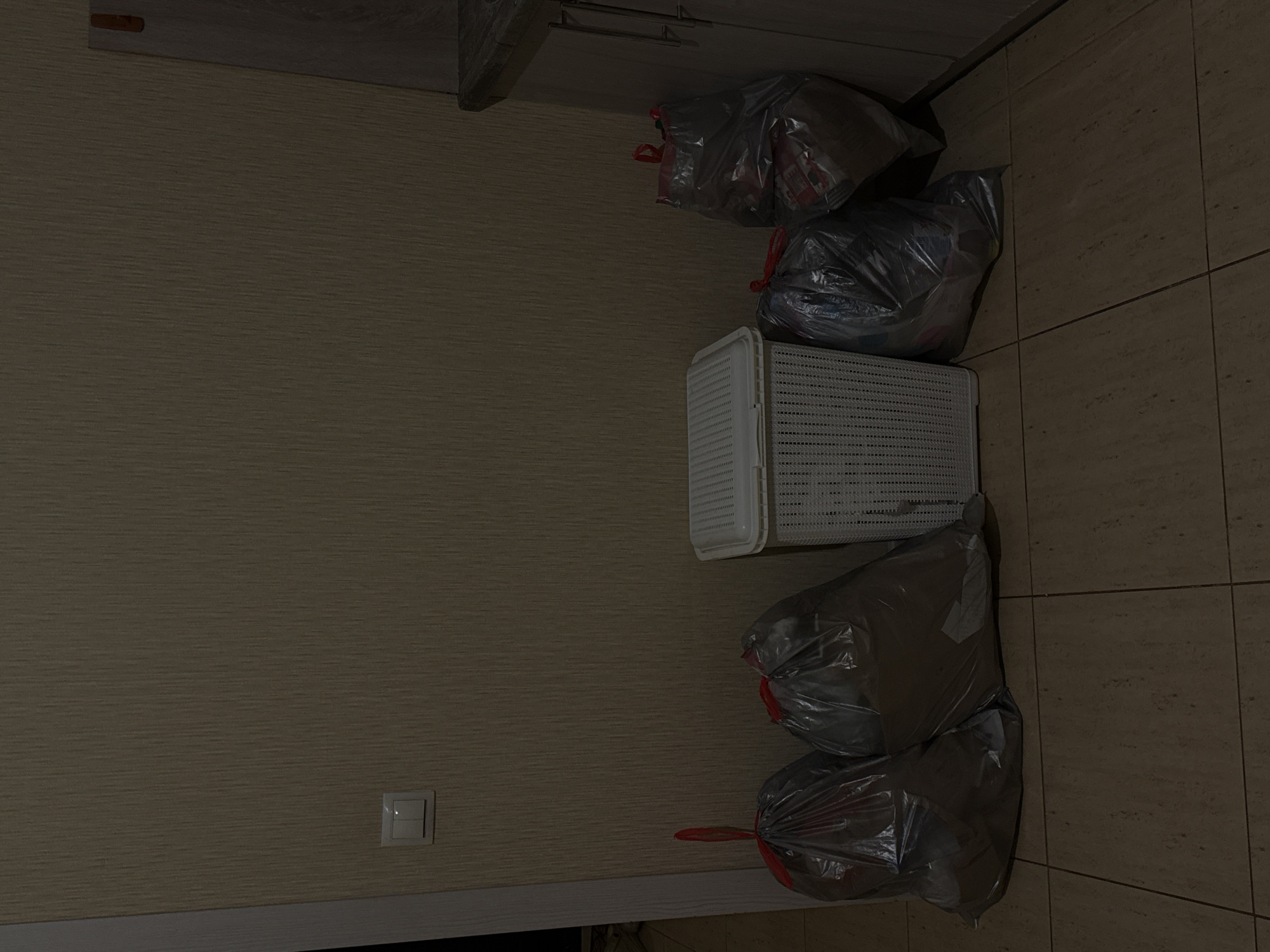 The image shows a dimly lit interior space, likely a kitchen or hallway, with tiled flooring. Against a textured, light brown wall are several objects. To the left, there is a light switch on the wall. In front of the wall, arranged from left to right, are five clear plastic trash bags, each tied with a red drawstring, and a white plastic laundry basket with a lattice pattern. The trash bags appear to be partially filled. To the far right, a portion of kitchen cabinetry with wooden doors and metallic handles is visible. The overall scene suggests a domestic environment where waste or laundry is being managed. There is no indication of the specific location in Abinsk, Russia.