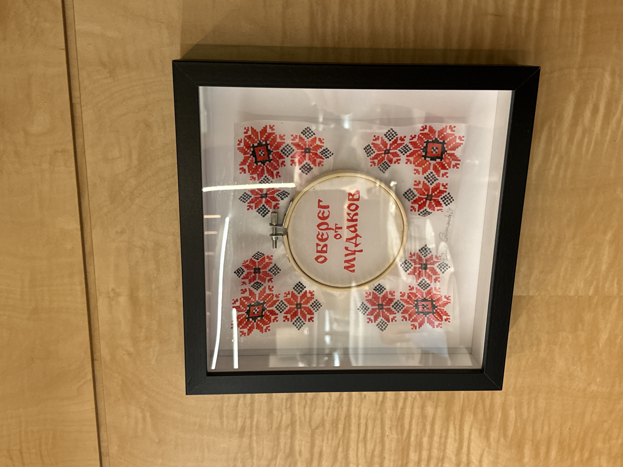 The image shows a framed piece of embroidery with a wooden hoop. The embroidery features red and black floral patterns on white fabric, and text in red lettering that reads "ОБЕРЕГ ОТ МУДАКОВ" (Charm against assholes). The frame is black and is hanging on a wooden wall. The overall scene suggests an art installation or a decorative piece in a home or public space. There is no identifiable context for its location within Moscow, Russia.