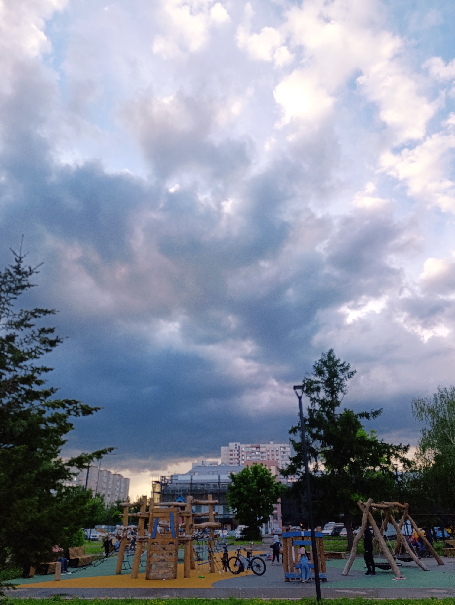 The image shows a playground in an urban setting. The sky is filled with dark, heavy clouds. In the background, there are apartment buildings, with one appearing to be under construction. The playground features wooden structures, a climbing frame, and swings, with people present. Bicycles are also visible. Trees frame the scene on either side. The playground appears to be located in Unknown, Unknown.