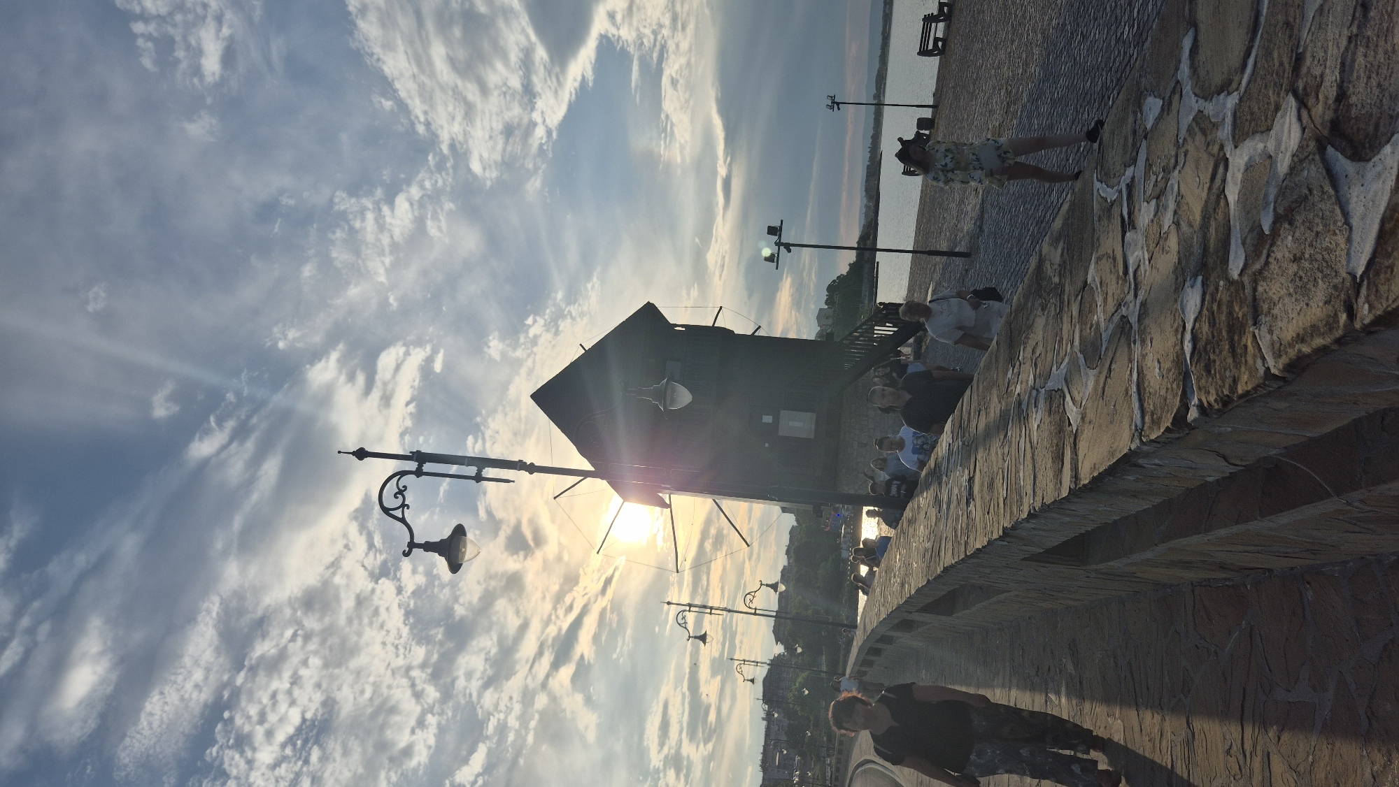 This is a medium-shot, vertical, outdoor photograph taken at a low angle of a promenade or walkway in Nesebar, Bulgaria. The sun is setting, casting long shadows and creating lens flare that obscures parts of the scene, especially around the central, dark wooden structure that appears to be a mill or tower. The sky is filled with scattered clouds, some illuminated by the setting sun.
In the foreground, a woman walks away from the camera on a paved walkway with a stone wall to her left. She is wearing a dark top and patterned pants. To her right, the stone wall has an uneven, mosaic-like pattern. Beyond the wall, more people are walking in the same direction. One woman in a floral dress and hat is closer to the camera on the right side of the walkway.
The dark wooden structure dominates the middle ground. It has a tall, steep roof and appears to have some sort of blades or wings, suggesting it's a windmill. Several streetlights with decorative designs are visible along the walkway, as well as some modern lampposts with simple fixtures. The sea is visible to the right of the walkway, with a distant shoreline and buildings on the horizon. A park bench is also visible on the right. The overall scene conveys a sense of a pleasant evening stroll in a historic coastal town.