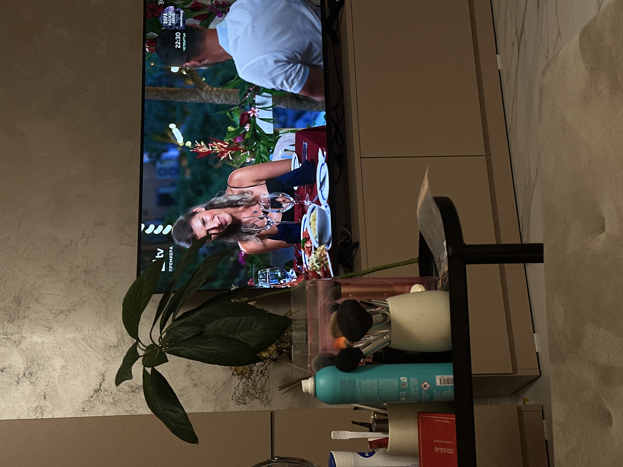 The image captures an indoor scene, likely a living room or bedroom, featuring a television displaying a program, and a shelf or table with personal items.
On the television screen, two individuals are shown having a meal or date in an outdoor, lush setting. A man, seen from behind, wears a light-colored polo shirt. A woman is seated at a table with drinks and food, smiling, and wearing a dark top. The background features tropical-like greenery and flowers. The screen displays Bulgarian text: "ВЯРА" (Faith), "ЛЮБОВ" (Love), "22:30" (10:30 PM), "ЧЕТВЪРТЪК" (Thursday), "ПРЕМИЕРА" (Premiere), and "ИЗБИРАНО" (Selected), along with "tv" and a channel logo, likely indicating a Bulgarian broadcast (consistent with the Sofia, Bulgaria context). The time suggests it's nighttime.
In the foreground, a large green leafy plant is visible. To the right of the TV, on a shelf unit, various personal items are organized. These include makeup brushes in a white mug and a clear container, a teal aerosol can (possibly dry shampoo or hairspray) with visible text and a flammable warning symbol, and a red box or book labeled "DOUGLAS GLAM FANTASY." Small dried flowers are also present. The surrounding walls have textured finishes in neutral tones.