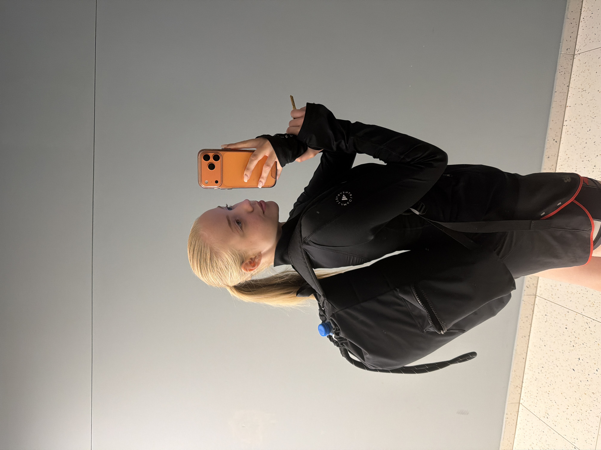 A young, light-skinned individual with blonde hair in a ponytail is taking a selfie in front of a large mirror. They are wearing a black long-sleeved athletic jacket with the logo of Adidas by Stella McCartney visible on the left chest. They also have on black shorts with a red trim. A black drawstring backpack with a blue water bottle is slung over their shoulder. They are holding an orange iPhone in their left hand and a small object resembling a wooden stick or key in their right hand. The background consists of a gray reflective surface (the mirror) and a tiled floor in the lower right corner. The location appears to be in an airport or a similarly modern, minimalist space in an unknown city.