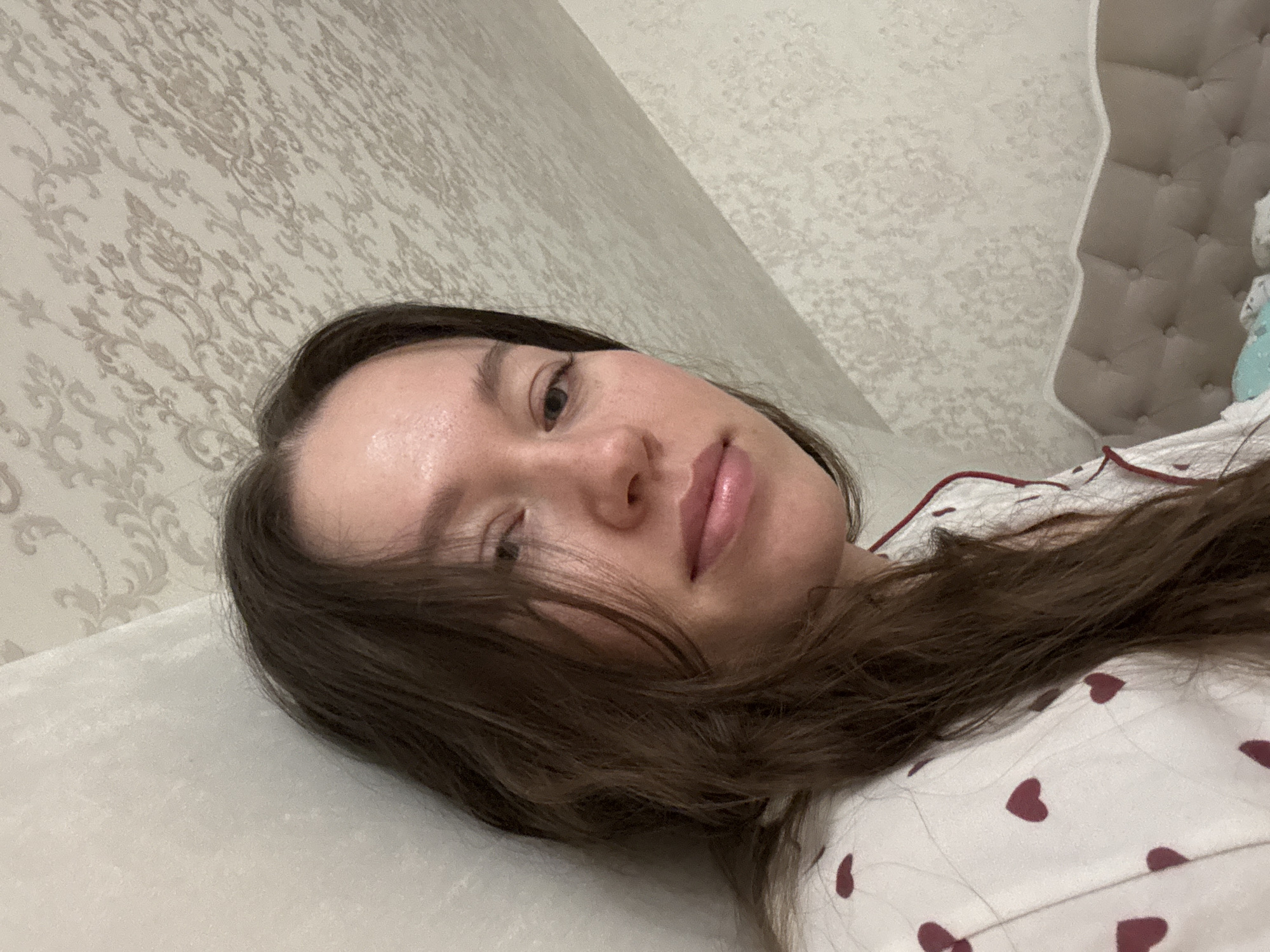 A woman with long, dark brown hair is lying down on a light-colored surface, possibly a bed, with her head tilted towards the camera. She is wearing a white top patterned with burgundy hearts. The background consists of a wall covered in light beige wallpaper with a floral or ornate pattern. Part of a padded headboard is visible in the upper right corner. The image appears to be taken indoors, possibly in a bedroom. There is no specific information connecting the image to the city of Ferma, Russia.