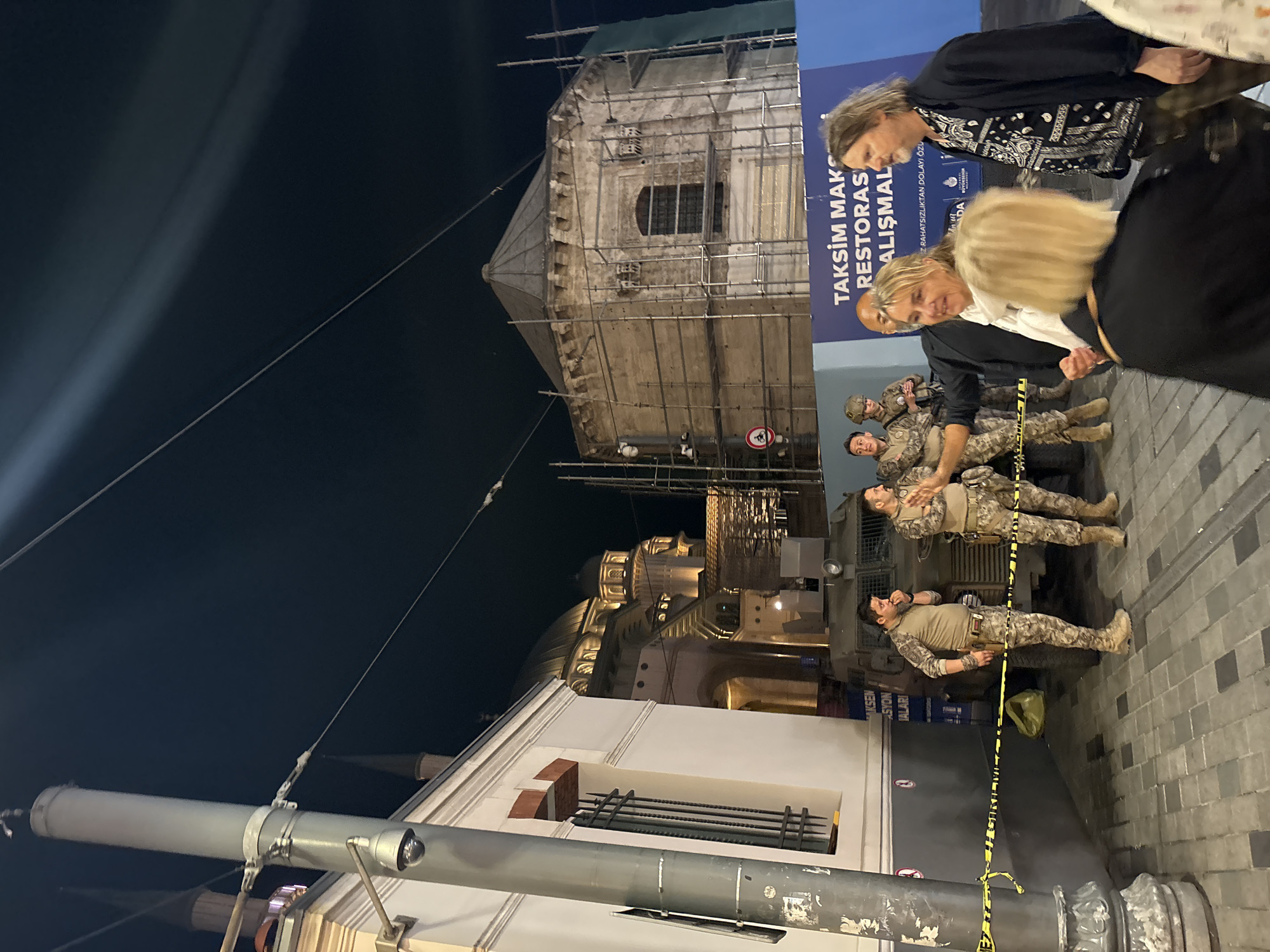 This nighttime image depicts an urban scene in Beyoğlu, Istanbul, Turkey, specifically near Taksim Square, as indicated by a partial blue banner reading "TAKSİM MAKS RESTORAS ÇALIŞMAL" (Taksim Max Restoration Works) and the Istanbul Metropolitan Municipality logo.
In the foreground, a tall, light grey utility pole stands on the left, equipped with a dome-shaped surveillance camera mounted low on its side. Yellow and black striped caution tape is stretched horizontally from this pole towards the right, delineating an area. The ground is paved with dark and light grey square and rectangular pavers.
To the right of the utility pole and behind the caution tape, a group of four men in camouflage uniforms and tactical gear are visible. Two men are prominently positioned near a dark green armored vehicle parked in the mid-ground. One of these men, facing right, has his hand near his mouth. The other, facing mostly forward, appears to be gesturing with his hands. Behind them, two more uniformed individuals, one wearing a helmet, are partially obscured by the vehicle. The armored vehicle has visible headlights and equipment on its front. A crumpled light-colored bag is on the ground behind the caution tape, near the vehicle.
In the immediate right foreground, three civilians are present. A woman with blonde hair, wearing a white scarf and a black jacket, is smiling and extends her right hand towards the uniformed men. To her right, a man with long, wavy hair and a beard, wearing a patterned black shirt and a black jacket, looks towards her. In front of them, a third woman with blonde hair is seen from the back, wearing a black top.
In the background, on the left, a white building with a barred window is partially visible. Further back and to the right, a large mosque or religious building with multiple illuminated domes and a slender minaret-like structure is visible under the dark sky. To the right of this mosque, a multi-sided, domed building, identified as being under restoration by extensive scaffolding covering its exterior, stands prominently. A red circular sign indicating "no cycling" is affixed near the base of the scaffolded building. Overhead wires traverse the dark sky.