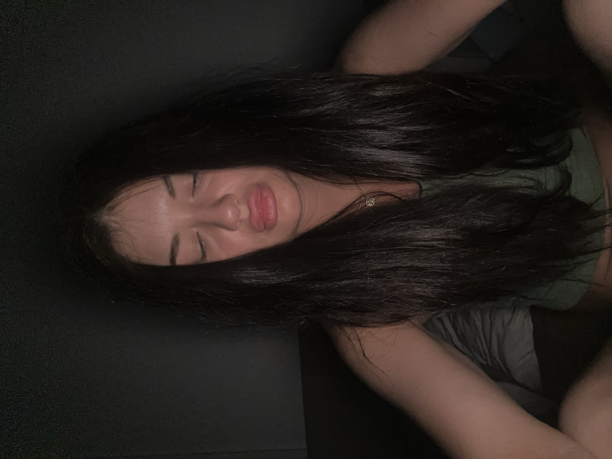 A woman with long, dark hair is lying down, eyes closed, with her arms visible. She appears to be on a dark-colored surface, possibly a bed or couch. She is wearing a green top, and a necklace is visible. The lighting is dim, and the location is not specified in the image. It is unclear what city the location is from.