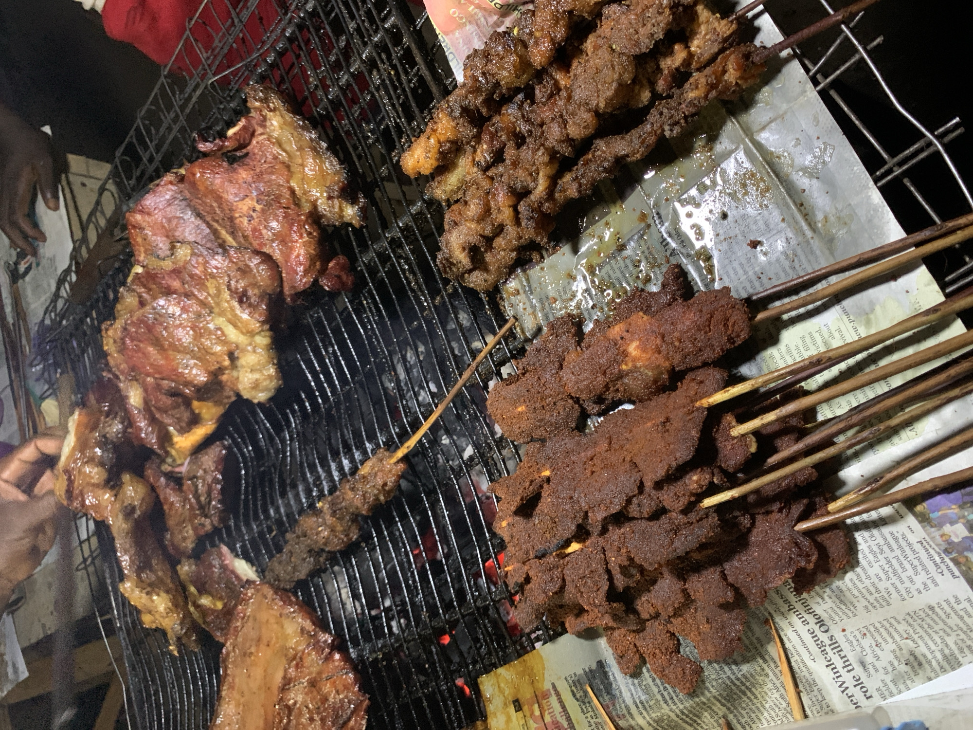 This image shows a close-up of food being grilled, likely on a roadside grill in Galadimawa, Nigeria, based on the presence of a newspaper, a common method to catch drippings and keep the grilling area clean. There are several types of grilled meat: large pieces of what appears to be beef or goat meat on the left, some skewers of smaller pieces of meat, and another set of skewers with a dark, spiced coating. A dark-skinned individual is present, possibly tending the grill. The grill itself is metal and has visible coals beneath. The environment is likely outdoors, possibly at night, considering the dark background.